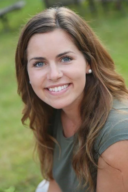 A young woman with long brown hair and blue eyes smiling outdoors, with a blurred green background.