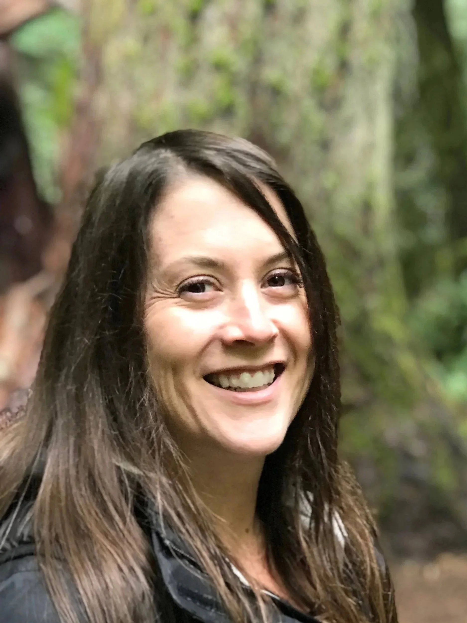 A smiling woman with long brown hair outdoors in a green, forested area.