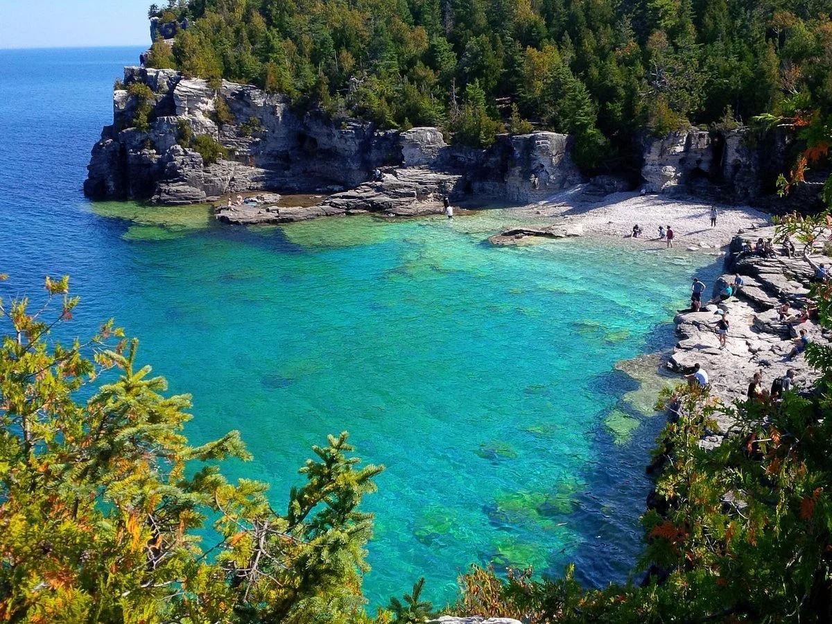A scenic coastal area with turquoise water, rocky shoreline, and dense green trees on cliffs, with several people relaxing and exploring along the shore.