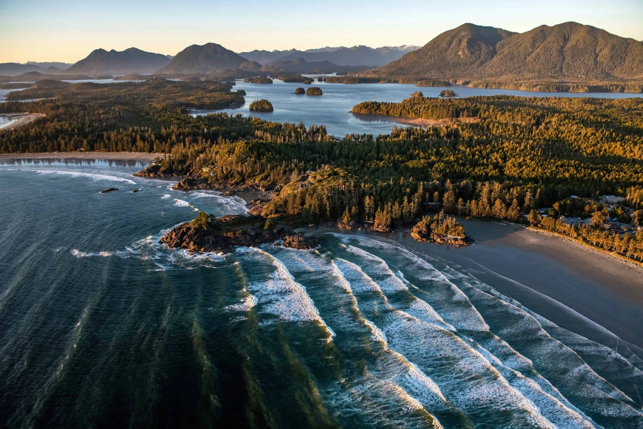 Aerial view of a coastline featuring waves crashing on the shore, dense forest, and distant mountains at sunset.