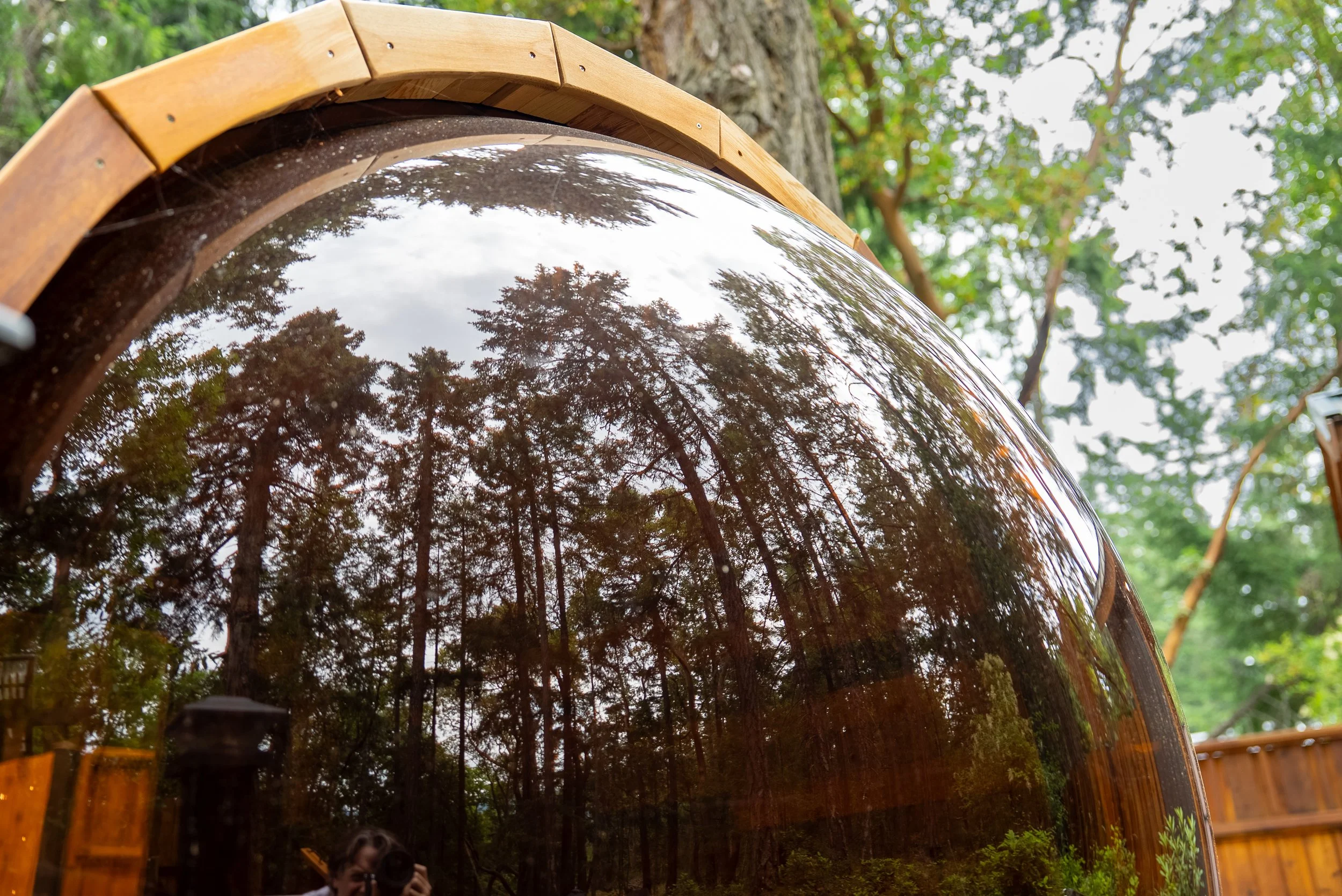 A round wooden sauna on a wooden deck in a forested backyard, with steps, plants, and string lights.