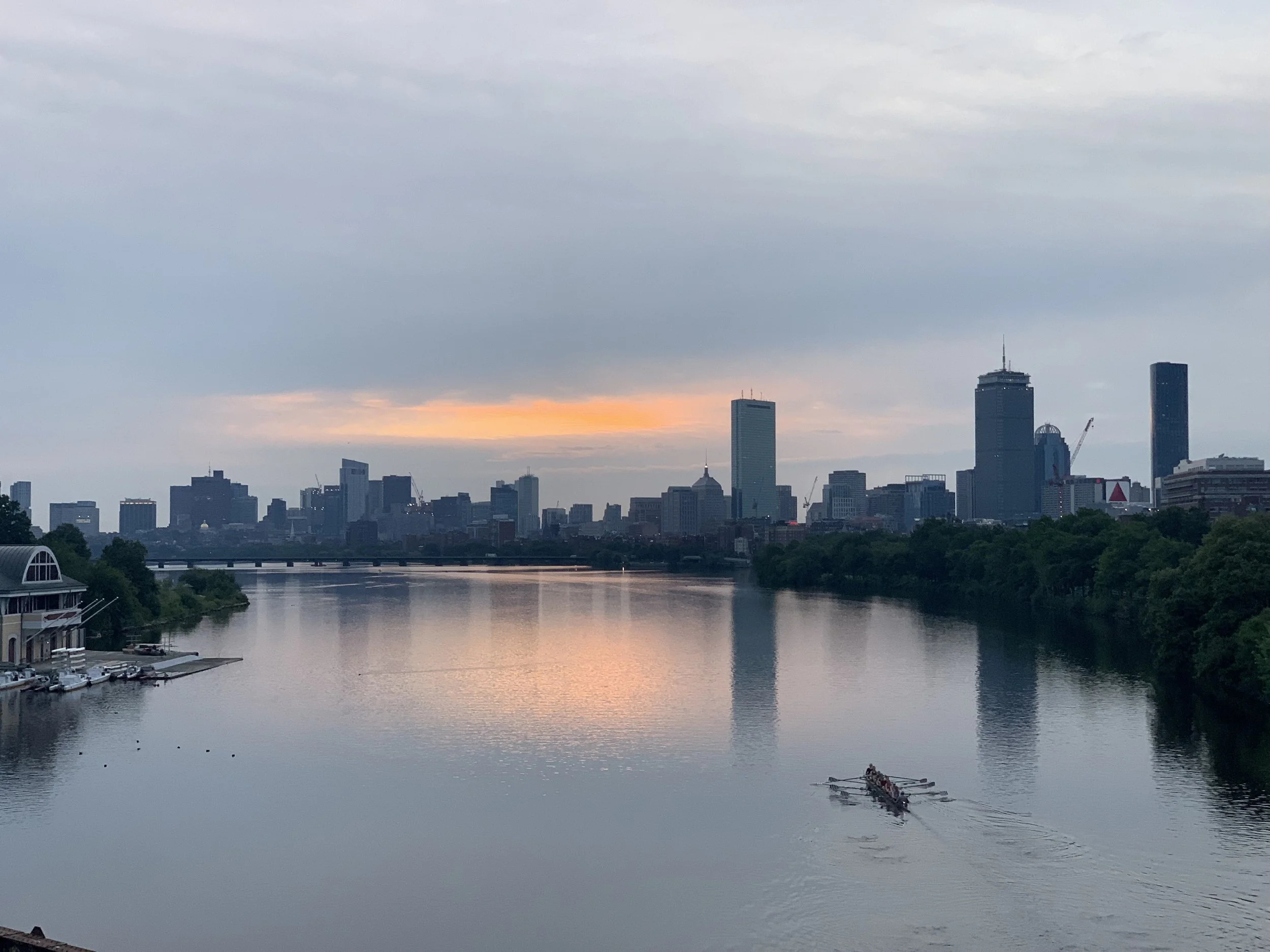 City skyline with tall buildings across a river at sunset, with a boat rowing in the water.