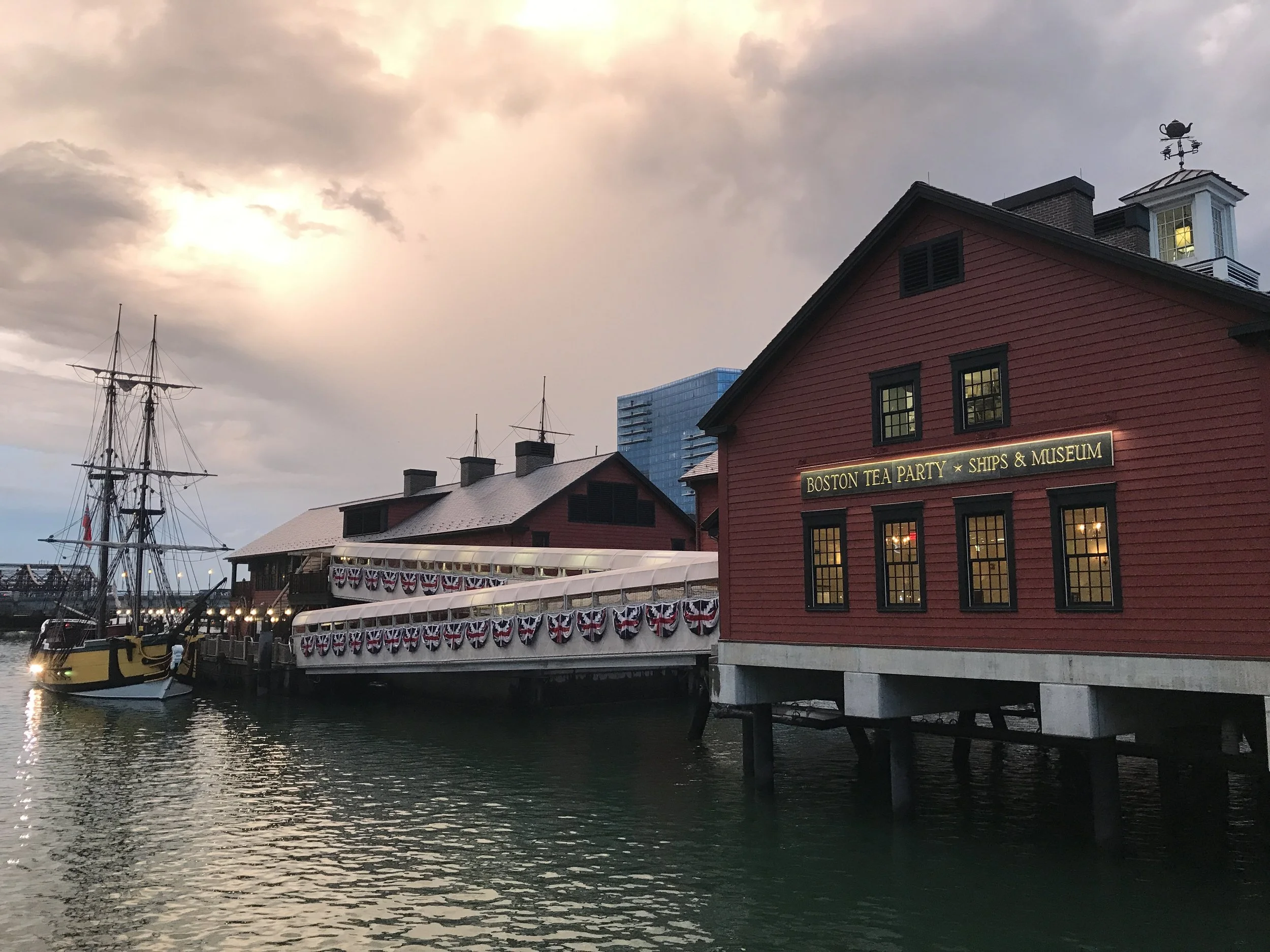 Vintage red building with a sign that reads 'Boston Tea Party, Ships & Museum' along a harbor at sunset, with a wooden ship and a small boat docked nearby, and patriotic bunting decorations.