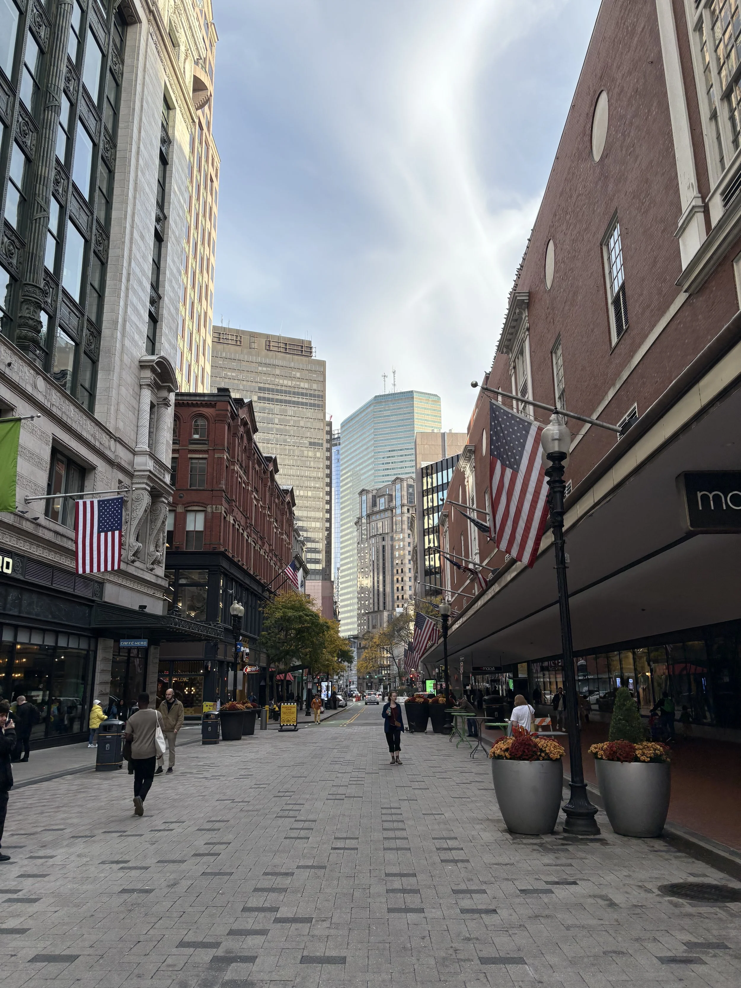 A city street scene with tall buildings, American flags hanging from street poles, and pedestrians walking along a paved sidewalk during daytime.
