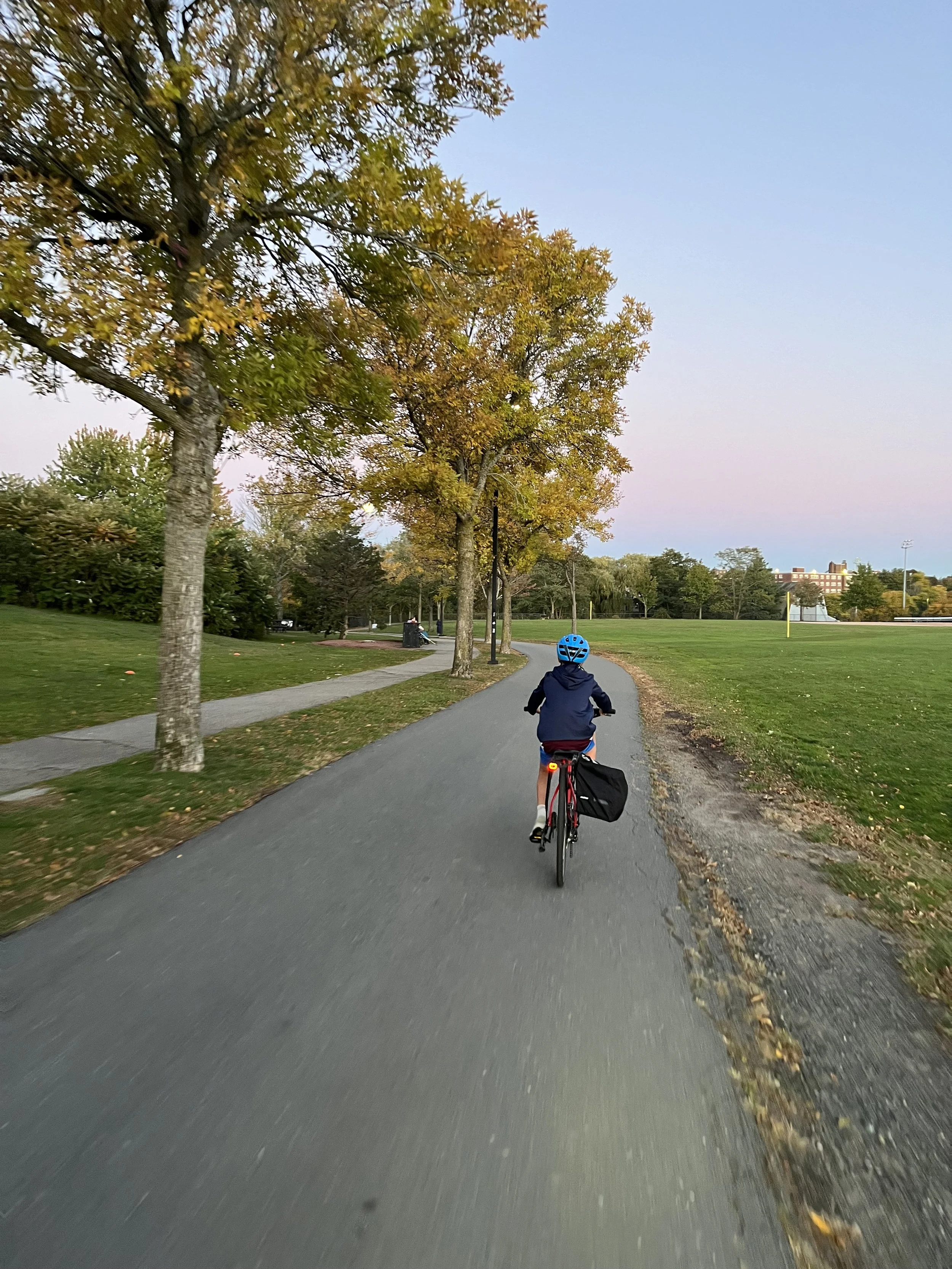 Child riding bicycle on a paved park trail with trees displaying fall foliage and a sports field in the distance.