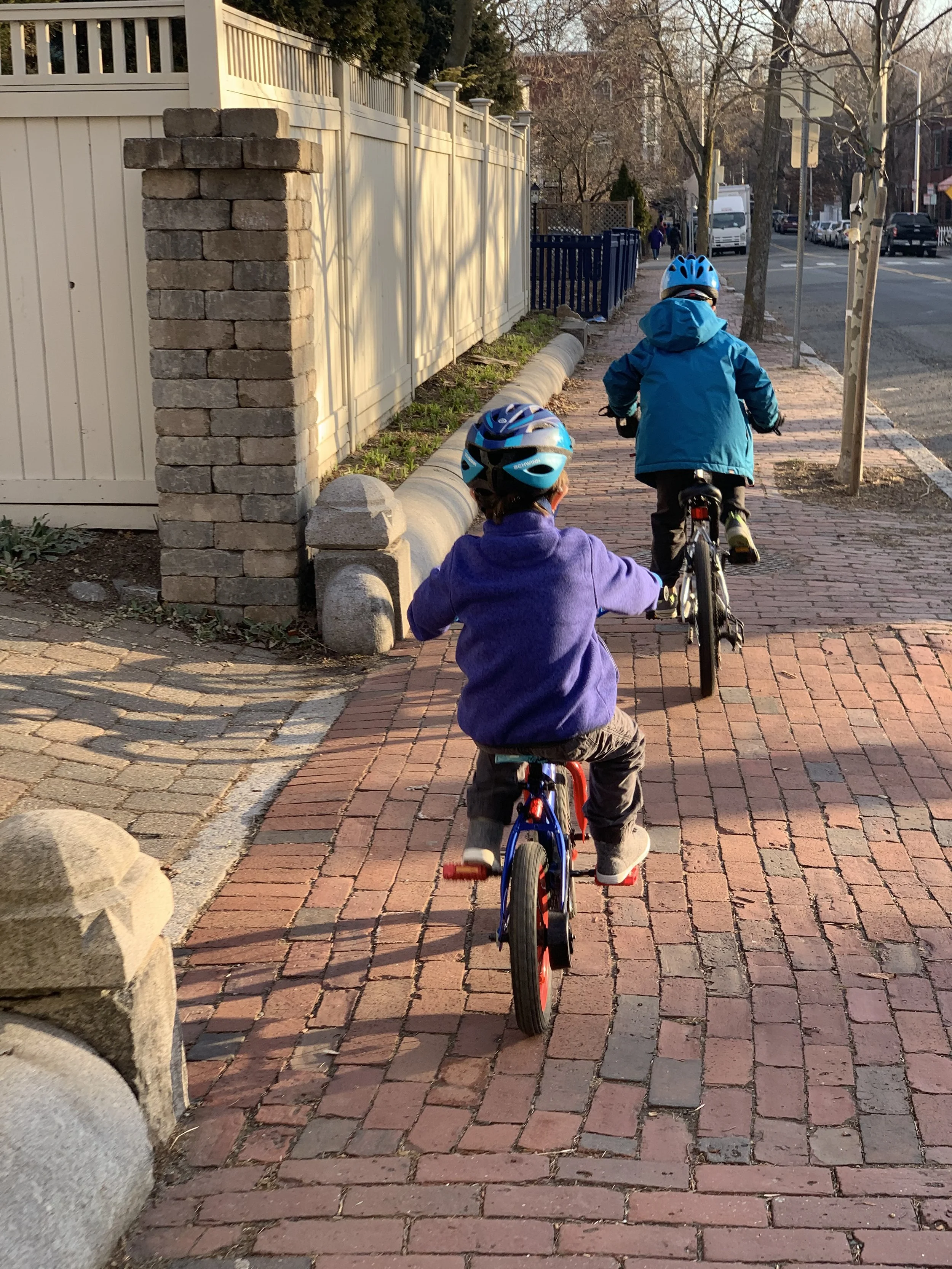 Two children riding bicycles on a brick sidewalk during late afternoon, wearing helmets and jackets, with a fence and trees on the side.