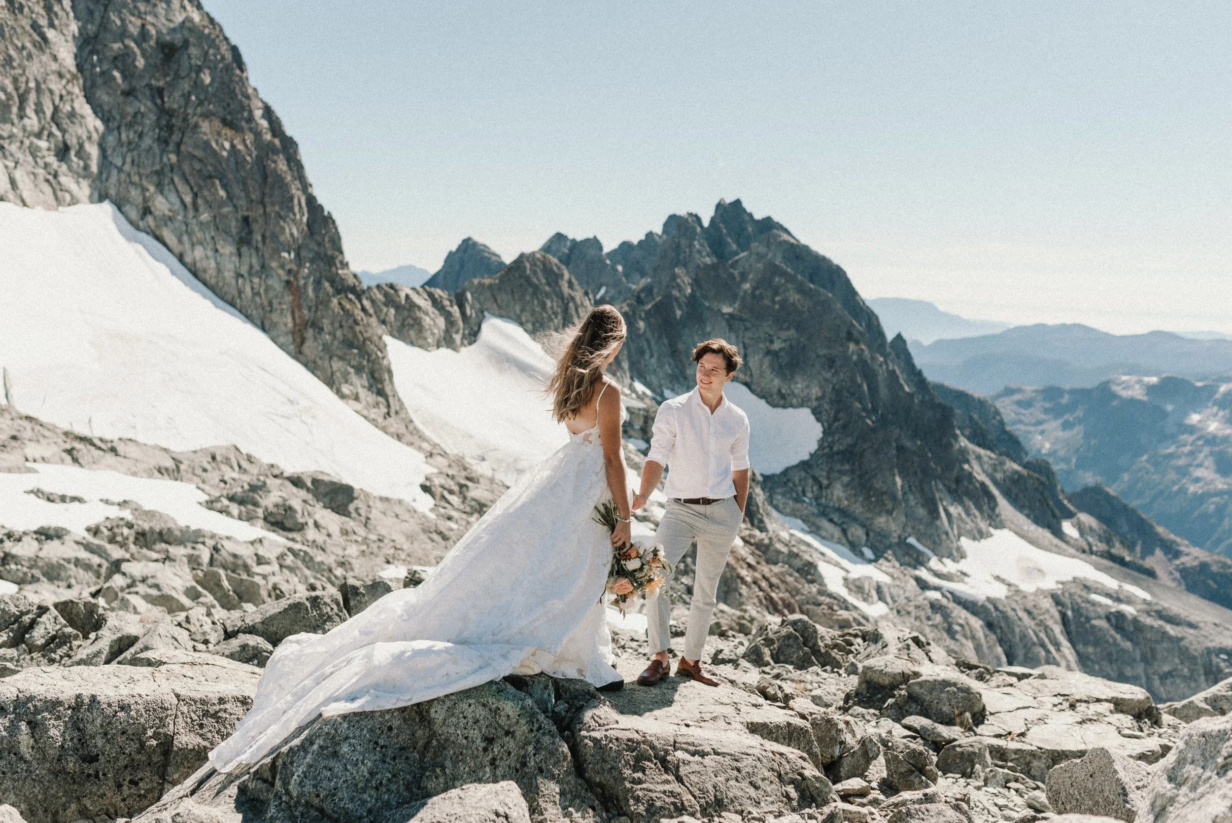 Bride and groom standing on a rocky, snowy mountain landscape during a wedding photoshoot, with rugged peaks in the background.