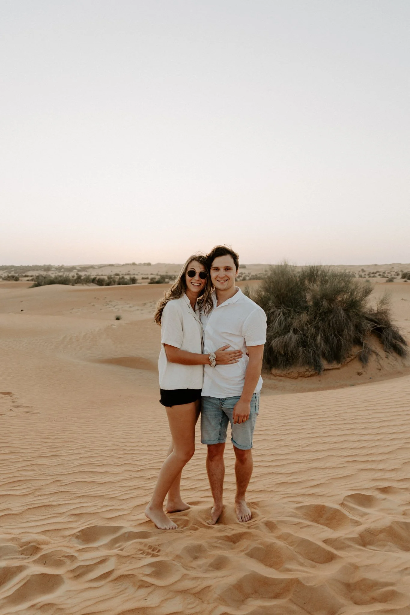 A couple standing barefoot on desert sand, embracing and smiling.