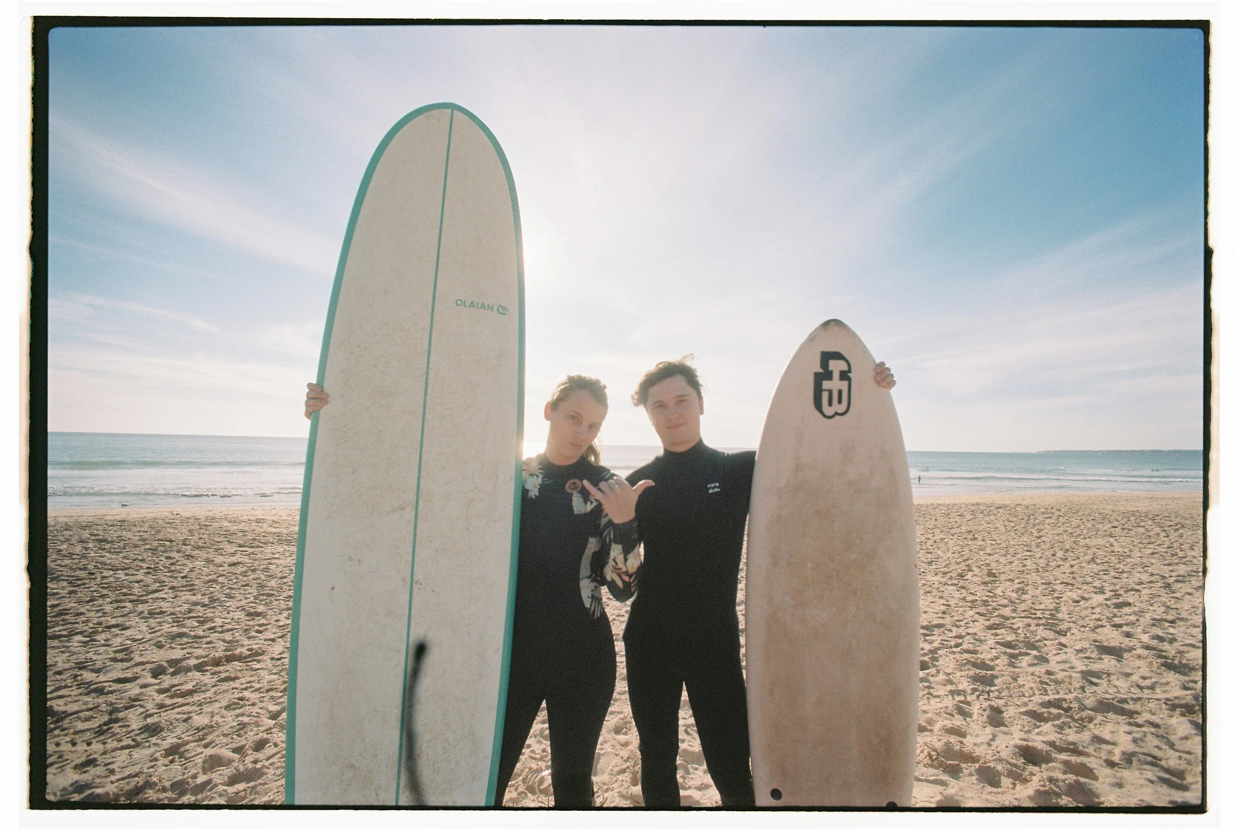 Two people in wetsuits holding surfboards on a beach with ocean in the background.