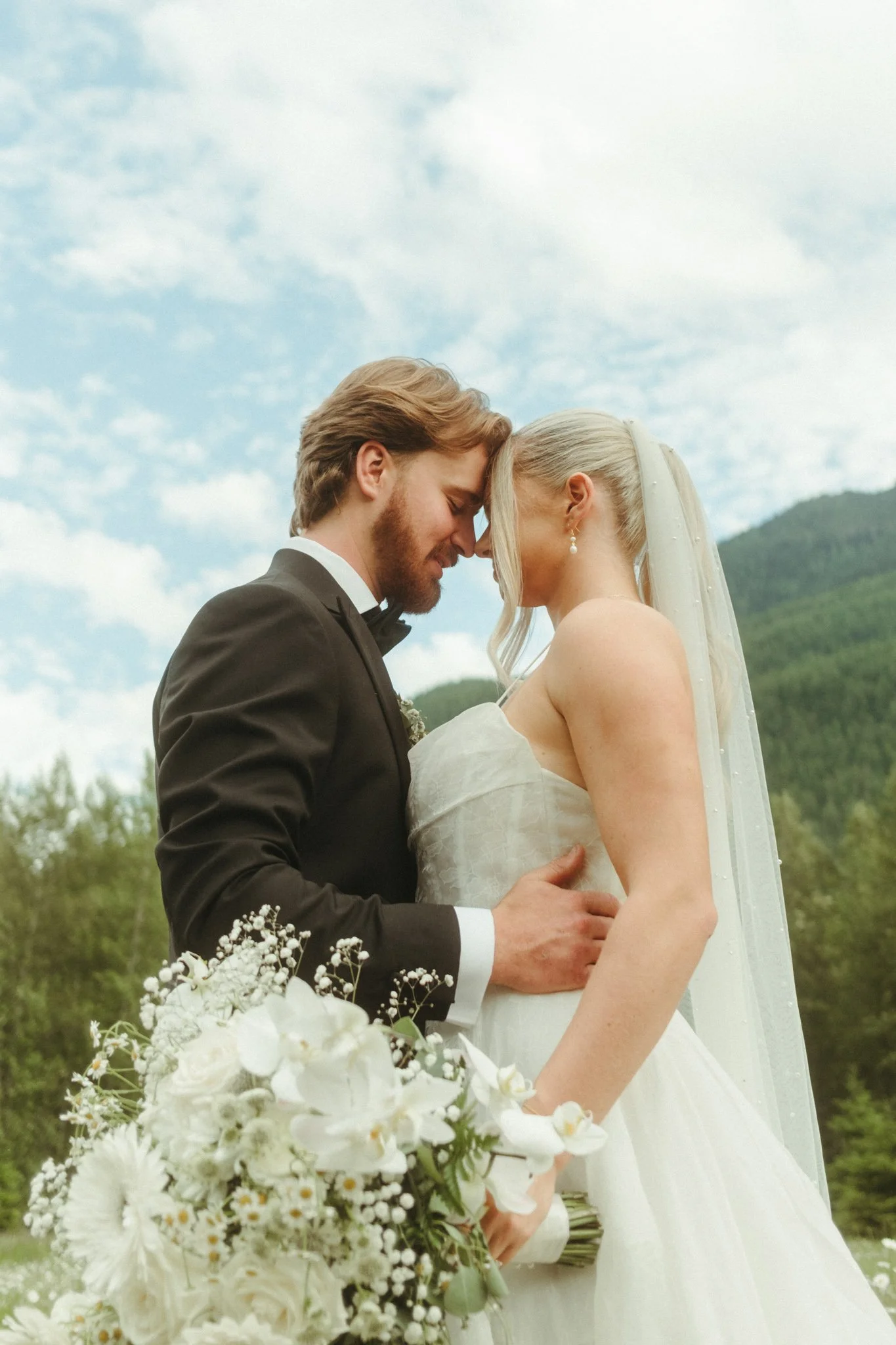 A bride and groom embracing outdoors with a bouquet of white flowers. The bride is wearing a white dress and veil, and the groom is in a black suit. Greenery and a mountain are in the background under a blue sky with clouds.