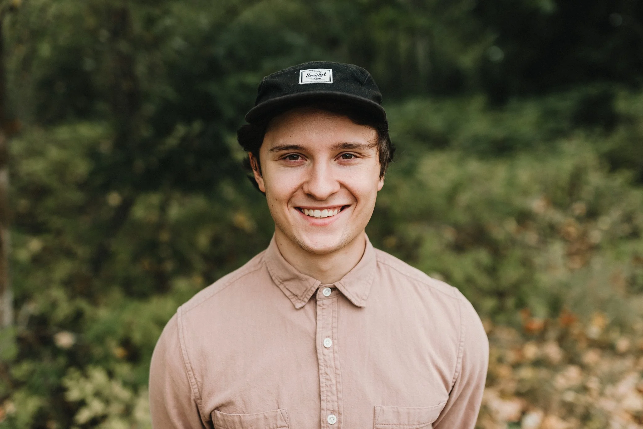 Smiling person wearing a black cap and beige shirt in an outdoor setting with greenery.
