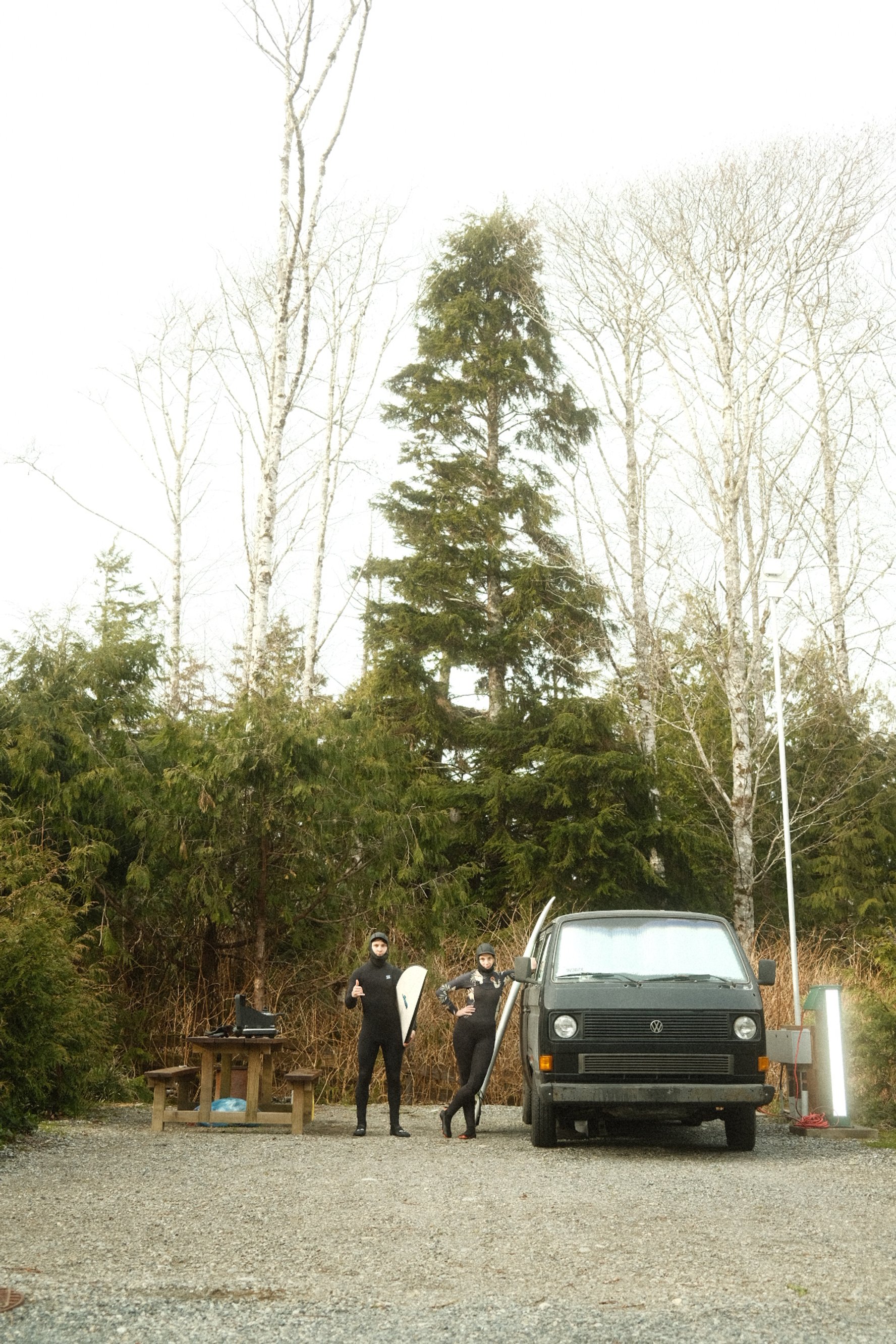 Two people in wetsuits standing next to a black van, one holding a surfboard, in a forested area with tall trees.