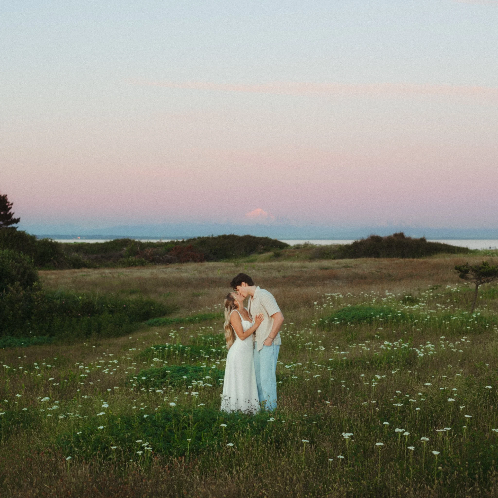 A couple embracing in a grassy field with wildflowers at sunset.