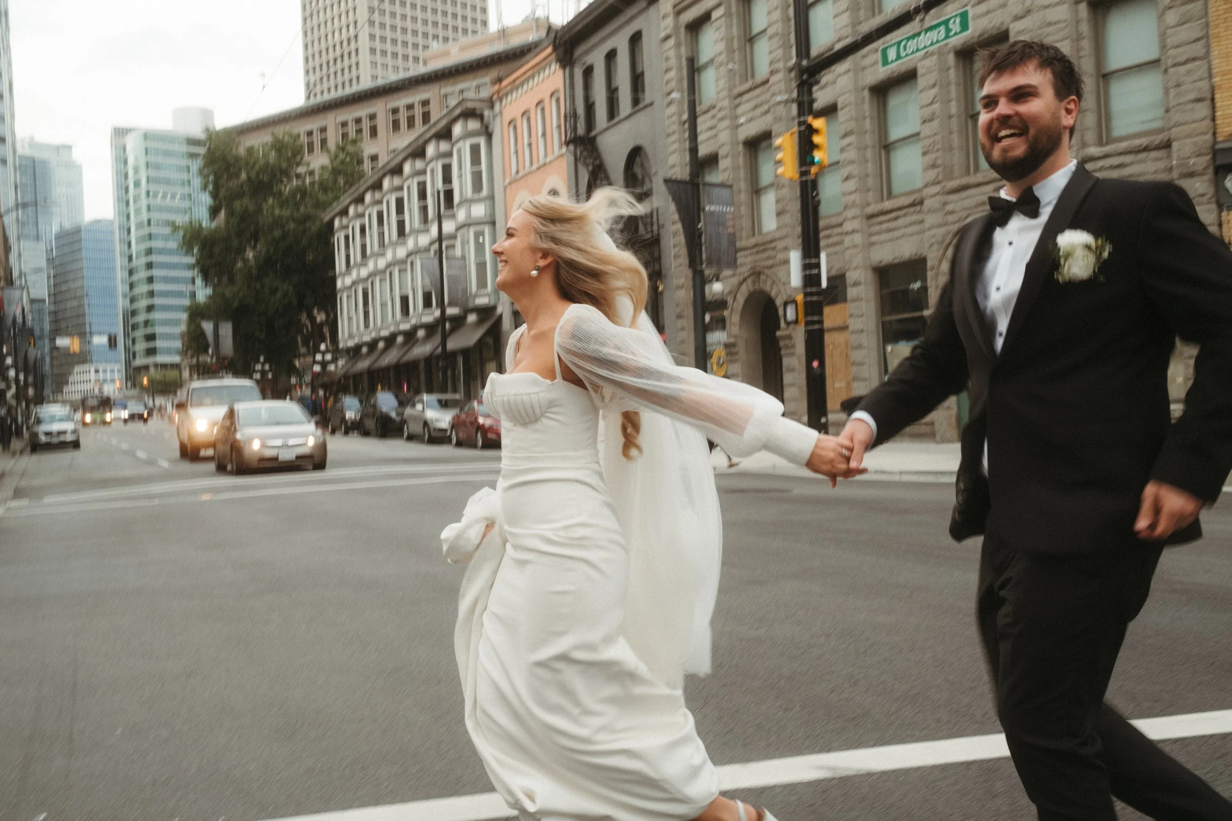 A bride and groom wearing wedding attire joyfully run across a street in an urban setting, with buildings and cars in the background.