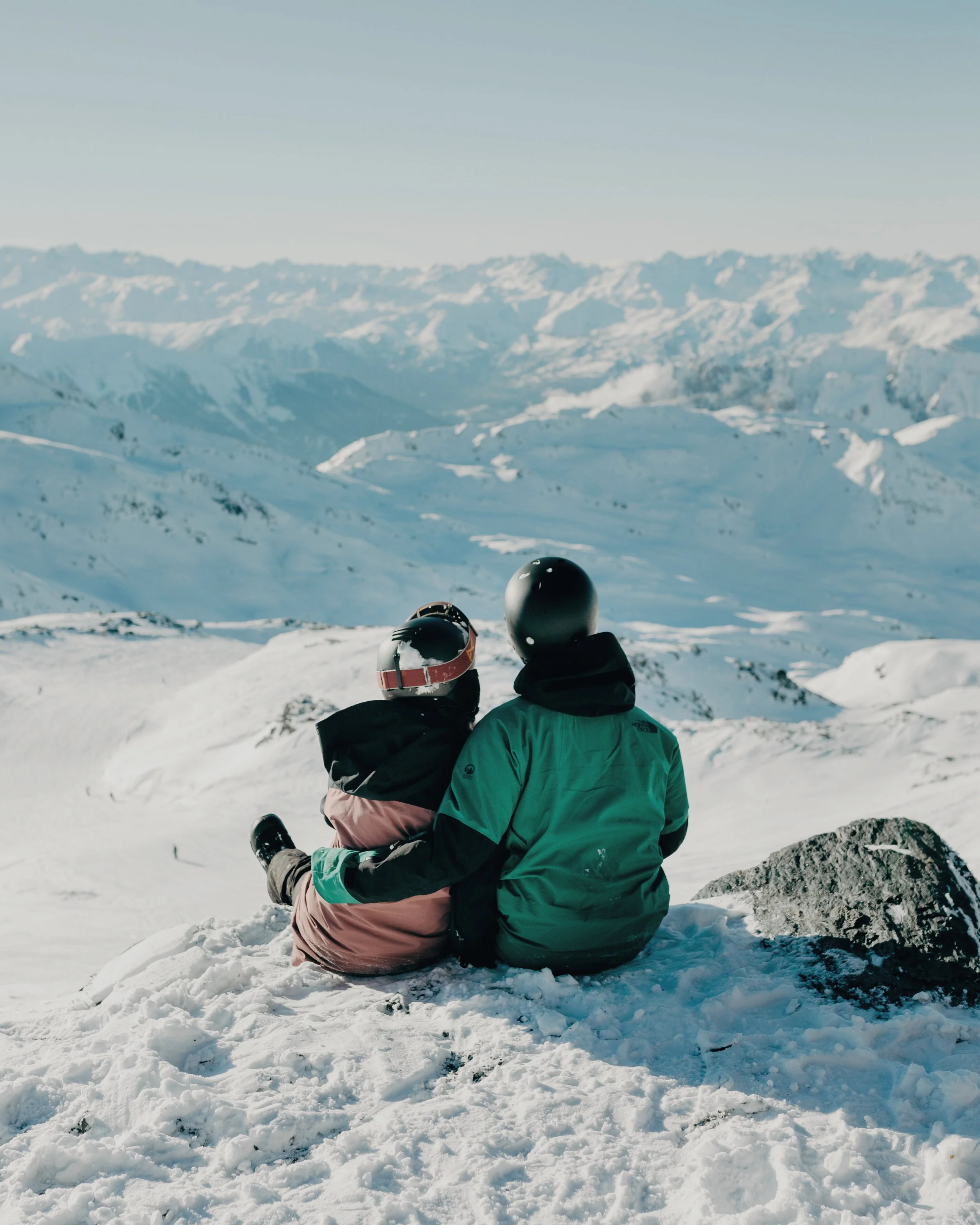 Two people in winter gear sitting on snow-covered ground, overlooking a mountainous landscape.
