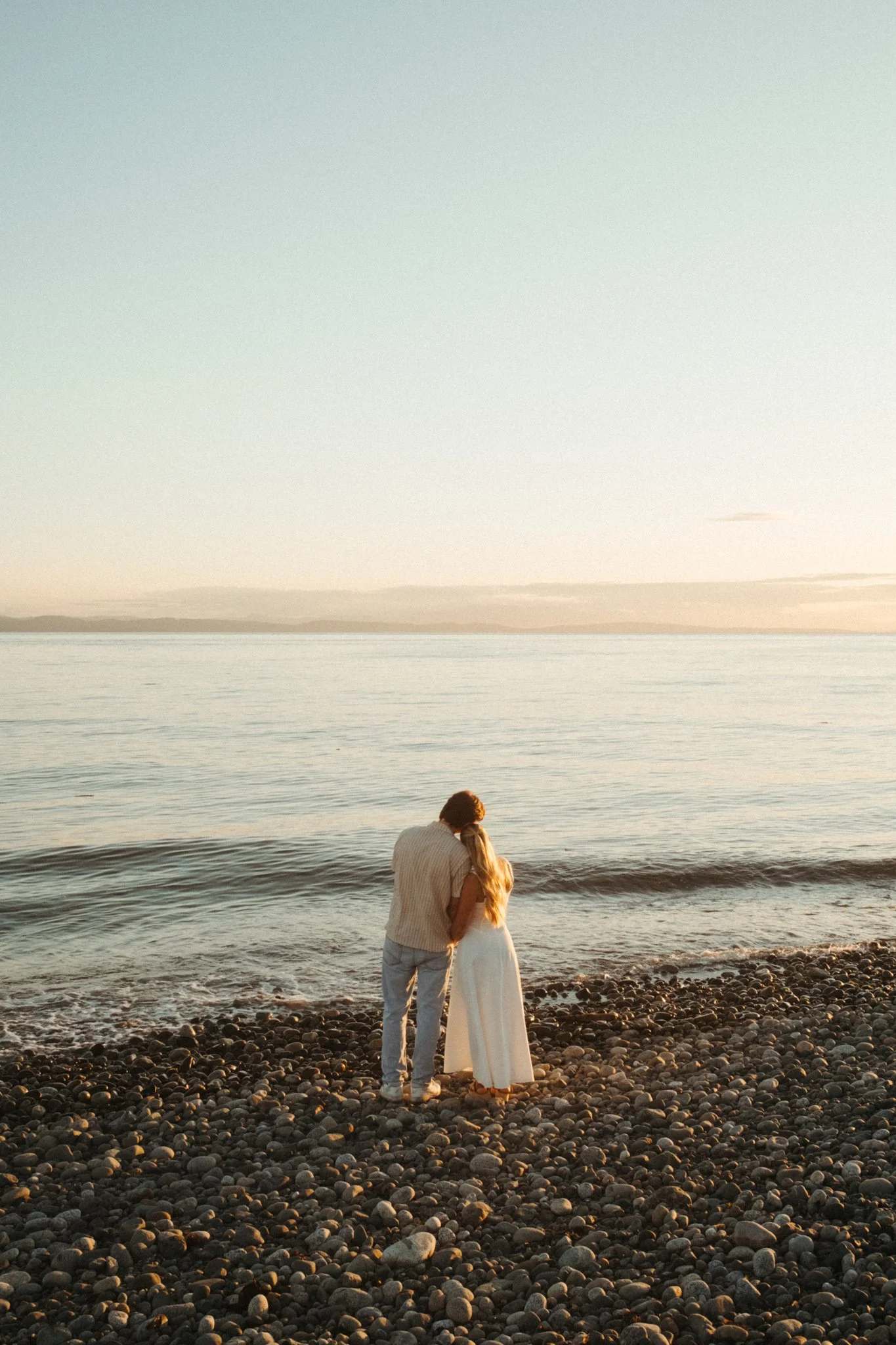 A couple embracing on a pebble beach at sunset, facing the ocean.