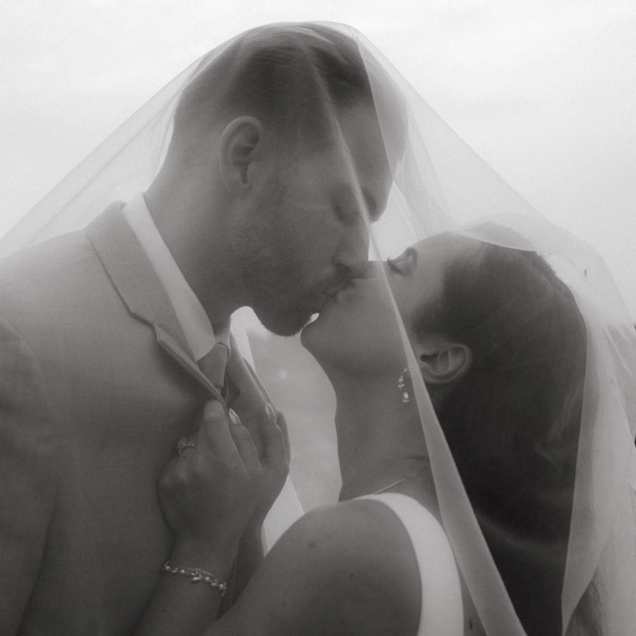 Black and white photo of a bride and groom kissing under a veil.