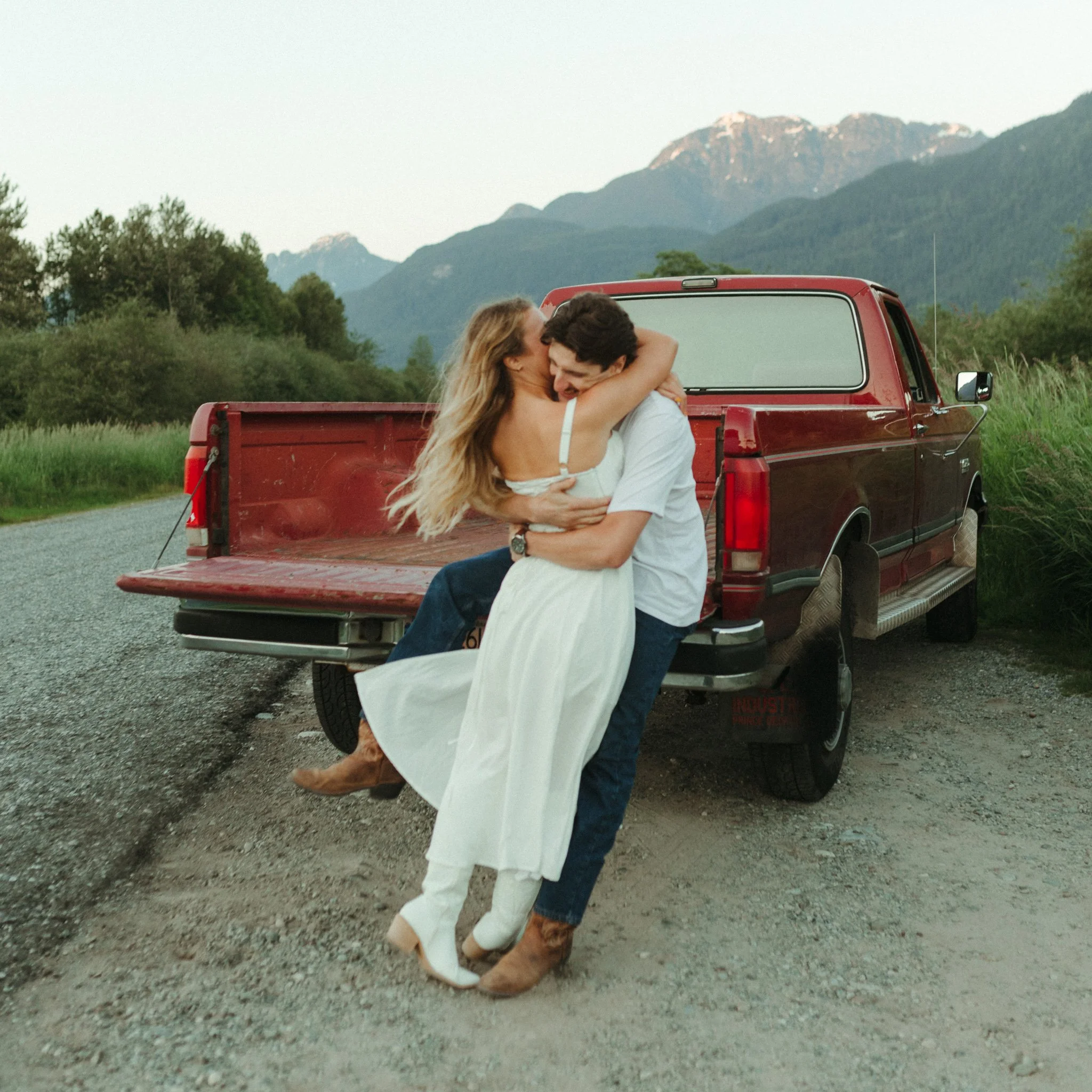 Couple embracing on the back of a red pickup truck parked on a gravel road with mountains in the background.