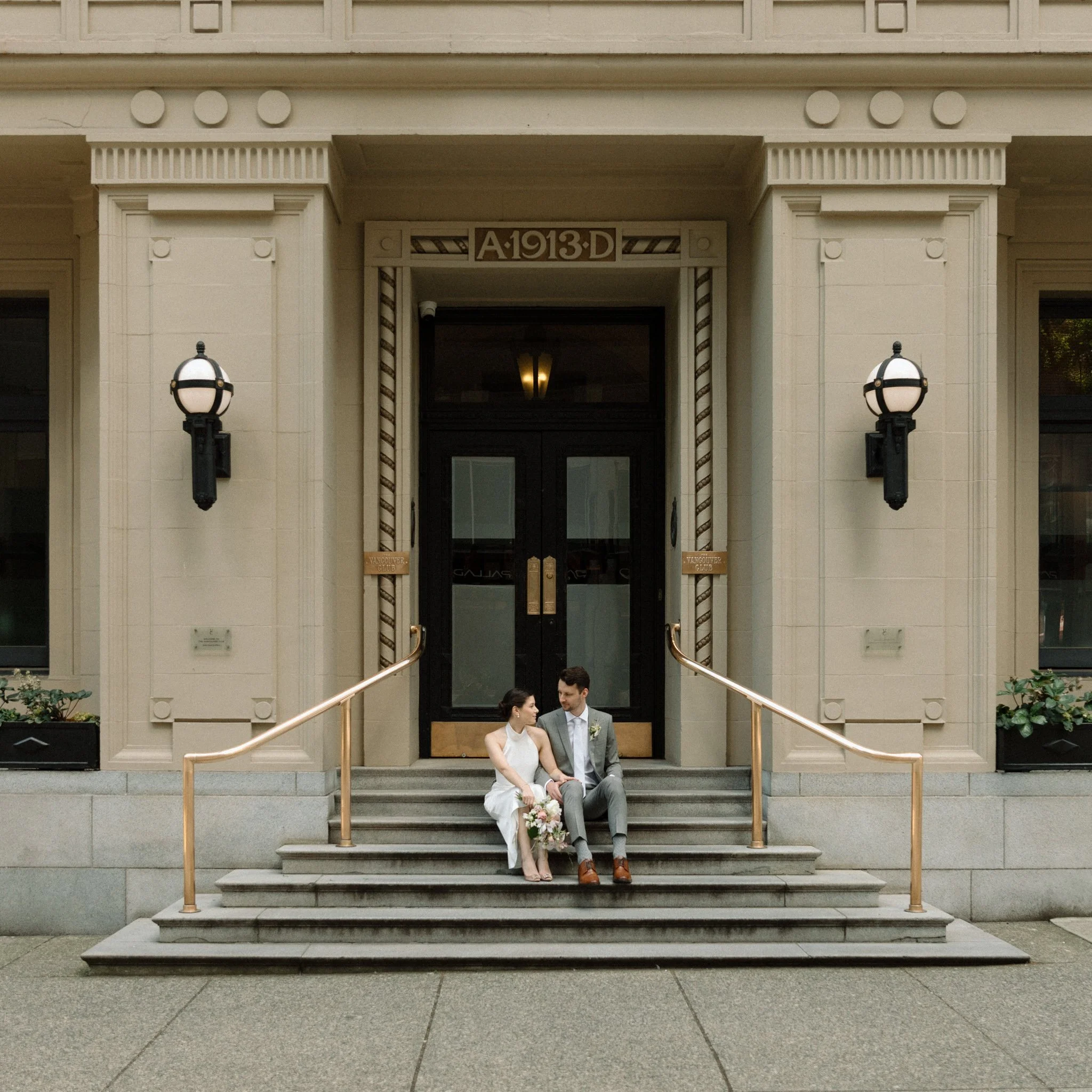 Bride and groom sitting on building steps, holding flowers, outside entrance marked "A 1913 D" with decorative lamps.