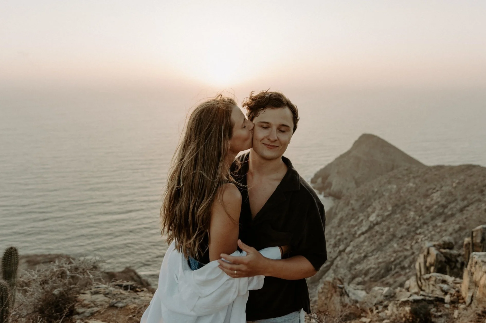 A woman kisses a man on the cheek at a rocky seaside cliff during sunset.