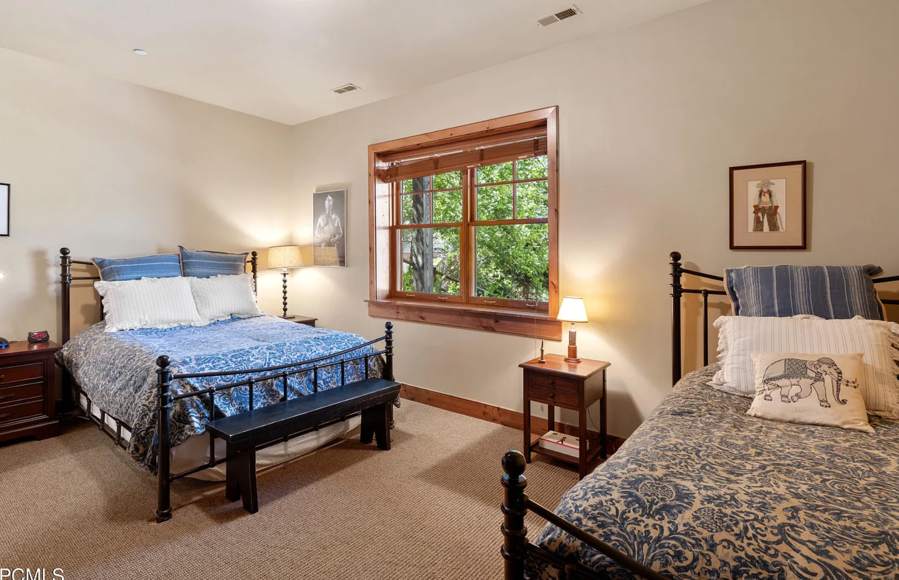Bedroom with two metal beds, blue patterned bedding, wooden window frame, nightstands, lamps, and wall art.