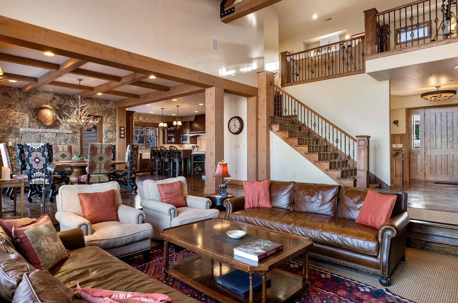 Spacious rustic living room with leather sofa, armchairs, wood-beamed ceiling, patterned rug, and decorative antler chandelier. Dining area and kitchen visible in the background with stone wall and upholstered chairs.