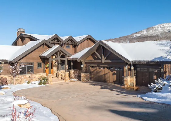 Large log cabin-style house with snow-covered roof and driveway, mountain backdrop, clear blue sky.
