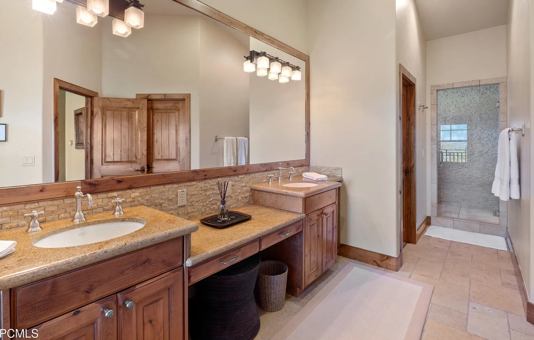 Modern bathroom with dual sinks, large mirror, wooden cabinets, glass shower, and beige tile flooring.
