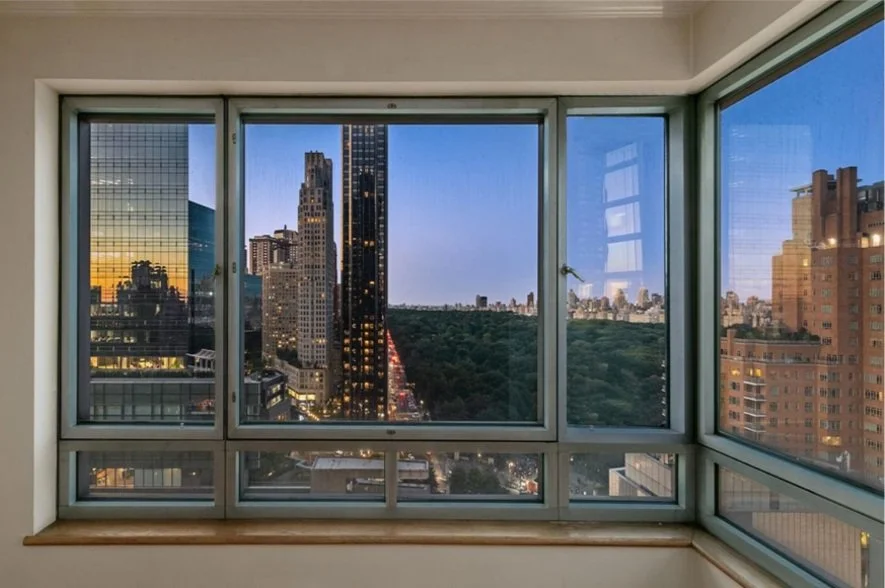 View of city skyline and Central Park through large window at twilight, with tall buildings on either side and a clear gradient sky.