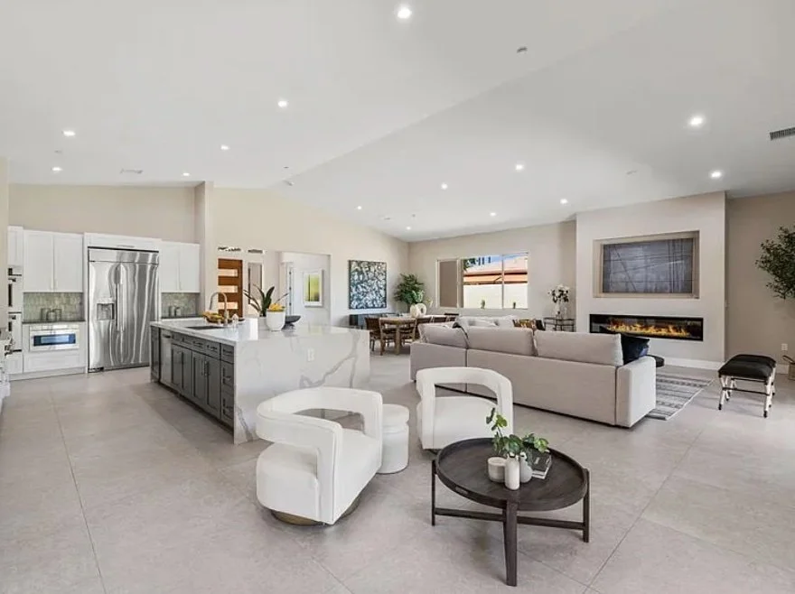 Modern open-concept living room with kitchen; white furnishings, large sofa, fireplace, round coffee table, stainless steel appliances, and minimalist decor.