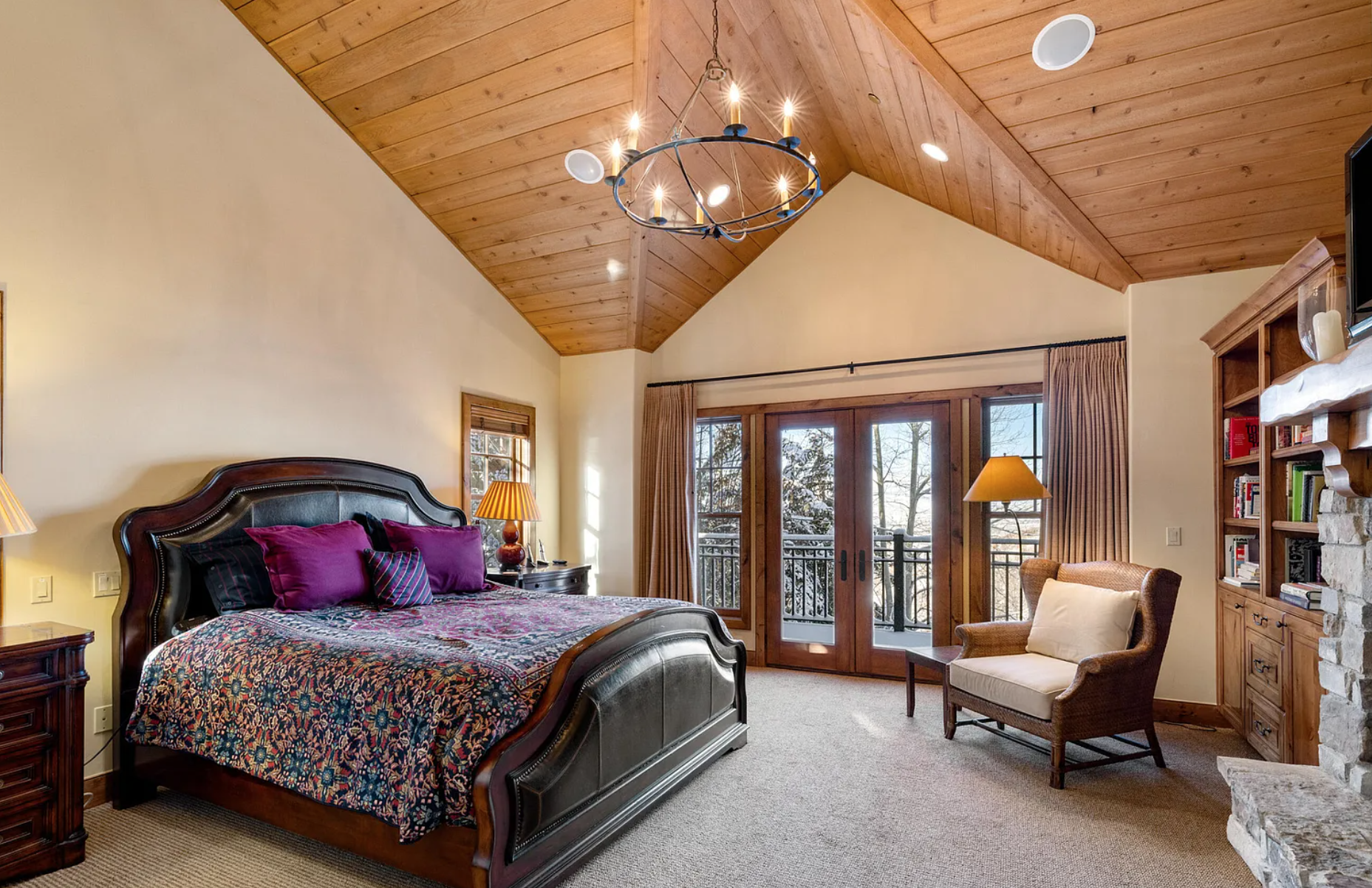 A cozy bedroom with vaulted wooden ceiling, large bed with colorful bedding, wood furniture including a bookshelf and dresser, armchair, chandelier, curtains, and glass double doors leading to a balcony.