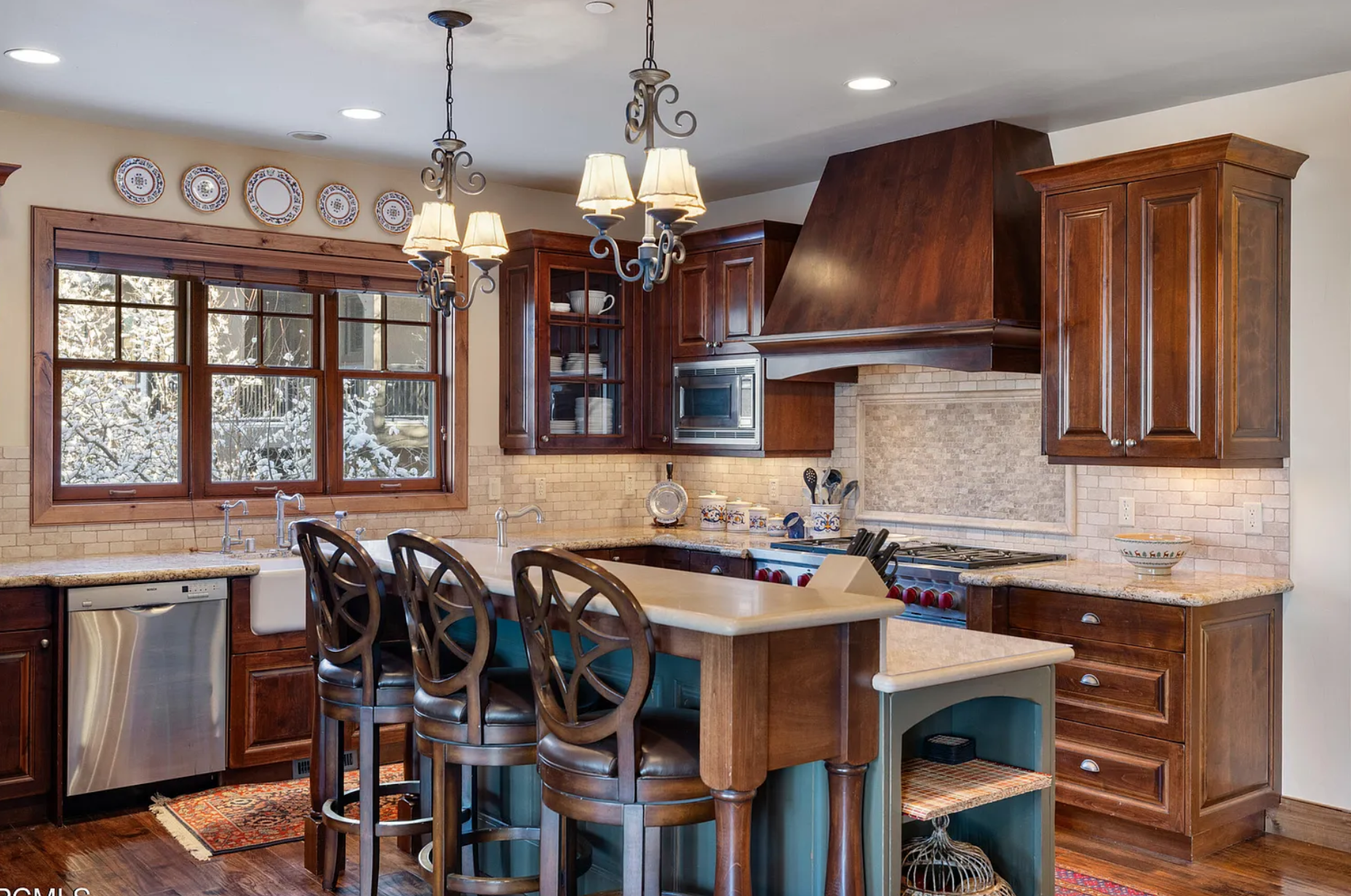Modern kitchen with wooden cabinets, granite countertops, and stainless steel appliances. Features a large island with bar stools, hanging pendant lights, and an oven range with a hood.