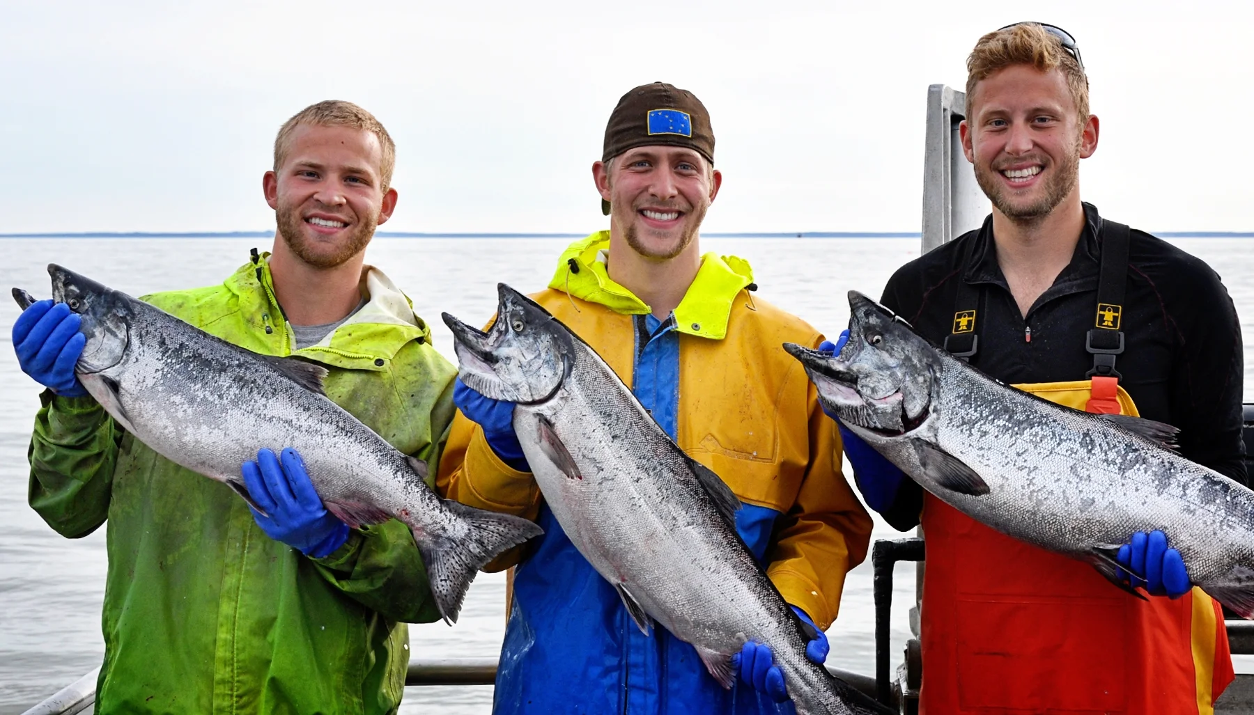 Three men holding large fish while on a boat, smiling, with water in the background.