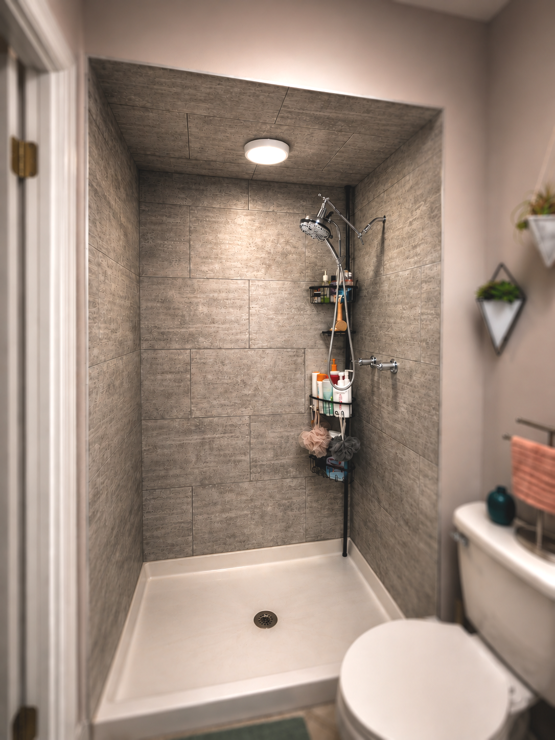 View of a small shower area with gray textured tiles, a ceiling light, a showerhead with a hanging shelf holding toiletries, and a drain on the white shower floor. Part of a white toilet and a corner of a wall with decorative small planters are visible.