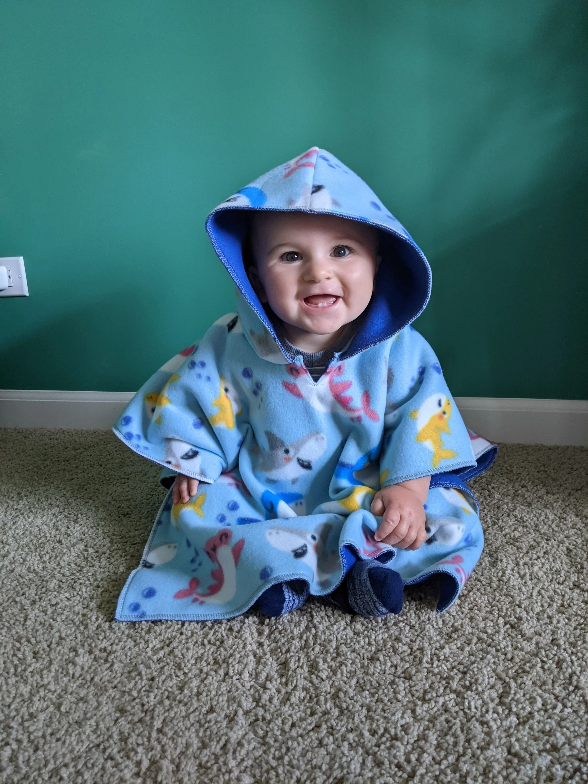 Smiling baby wearing a blue hooded blanket with cartoon animal prints, sitting on carpet in front of a green wall.