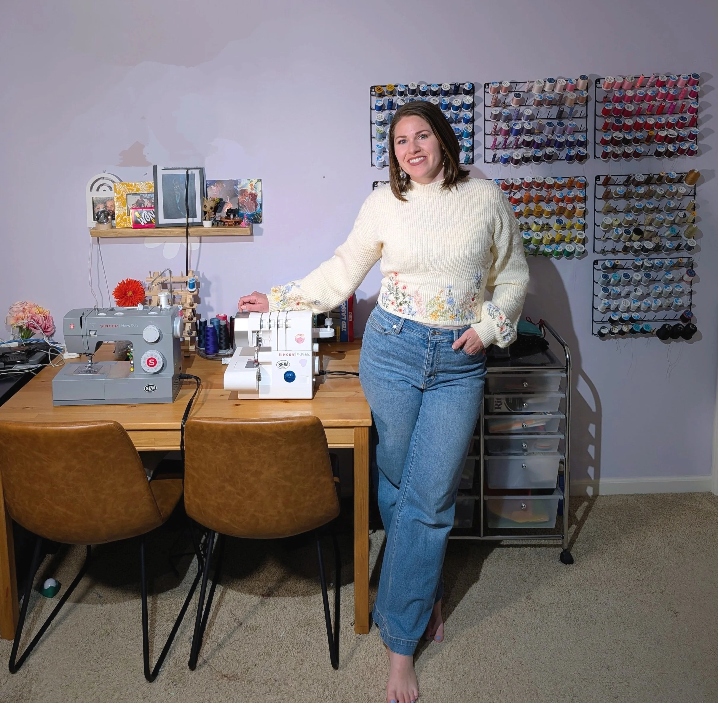 A woman standing beside a home sewing workspace with sewing machines, a wooden desk, and spools of thread on the wall.