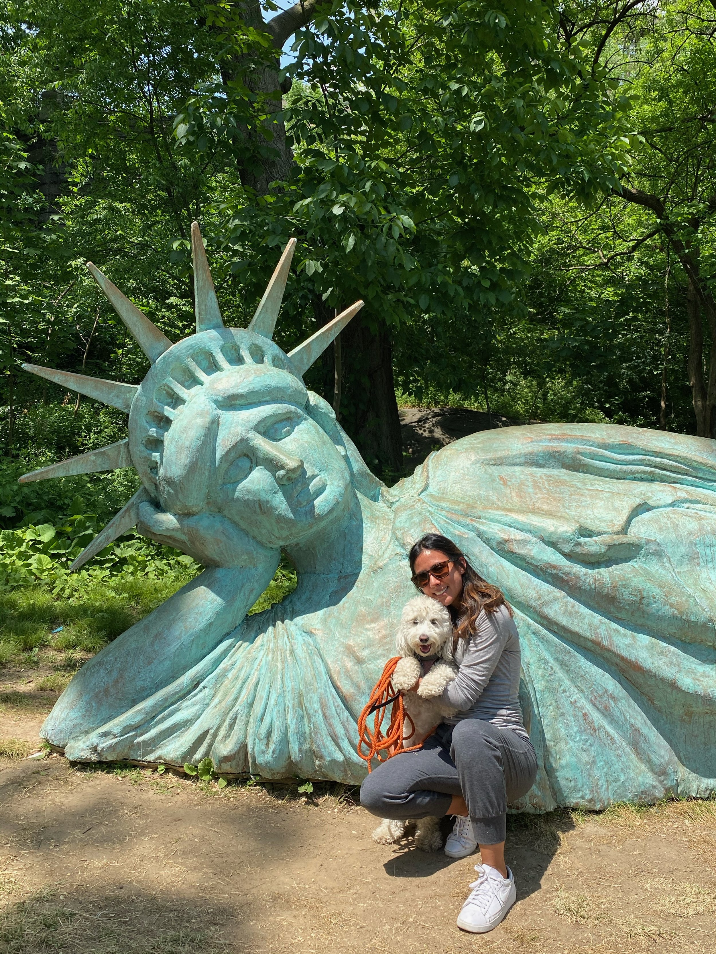 A woman with sunglasses crouching down and hugging a white fluffy dog, in front of a large sculpture of the Statue of Liberty in a green outdoor park.