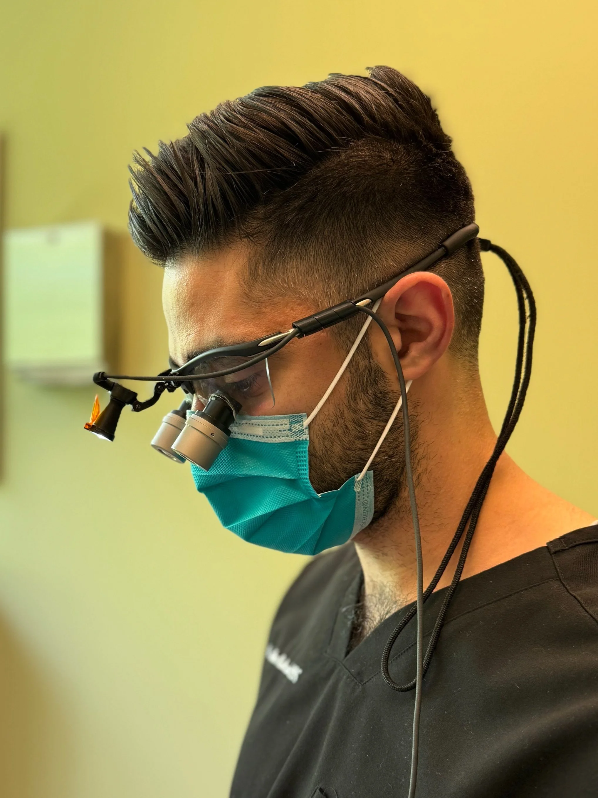 A man wearing a black medical scrub, a blue surgical mask, and magnifying loupes with a headlamp, looking downward.