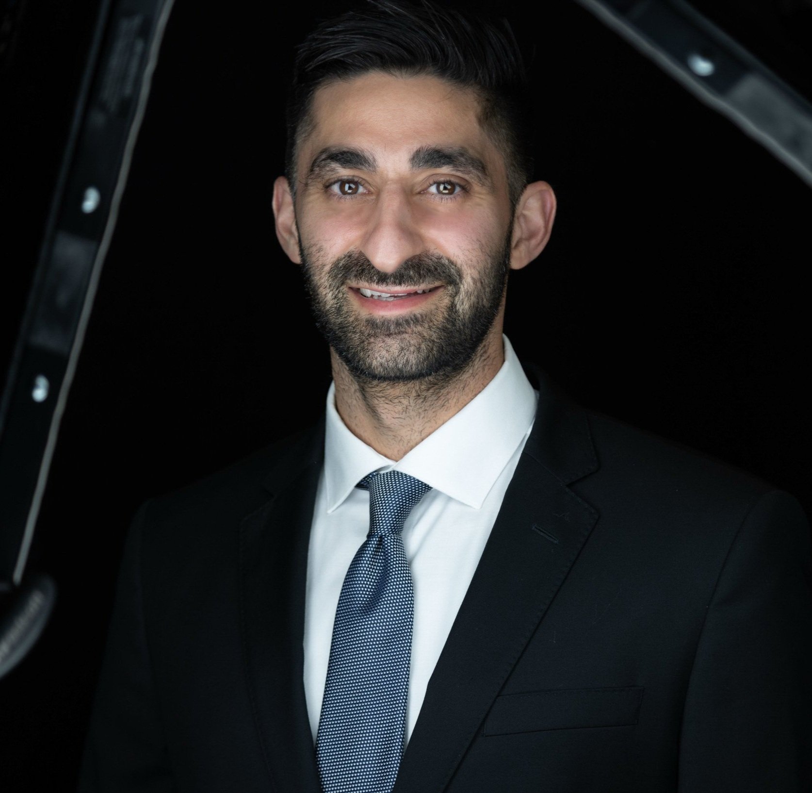 Portrait of a man in a black suit, white shirt, and patterned tie, smiling in front of a dark background.