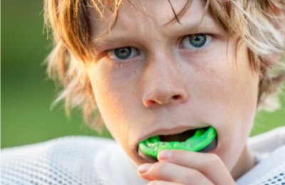 Close-up of a young boy with blond hair and blue eyes biting down on a green toy in his mouth.