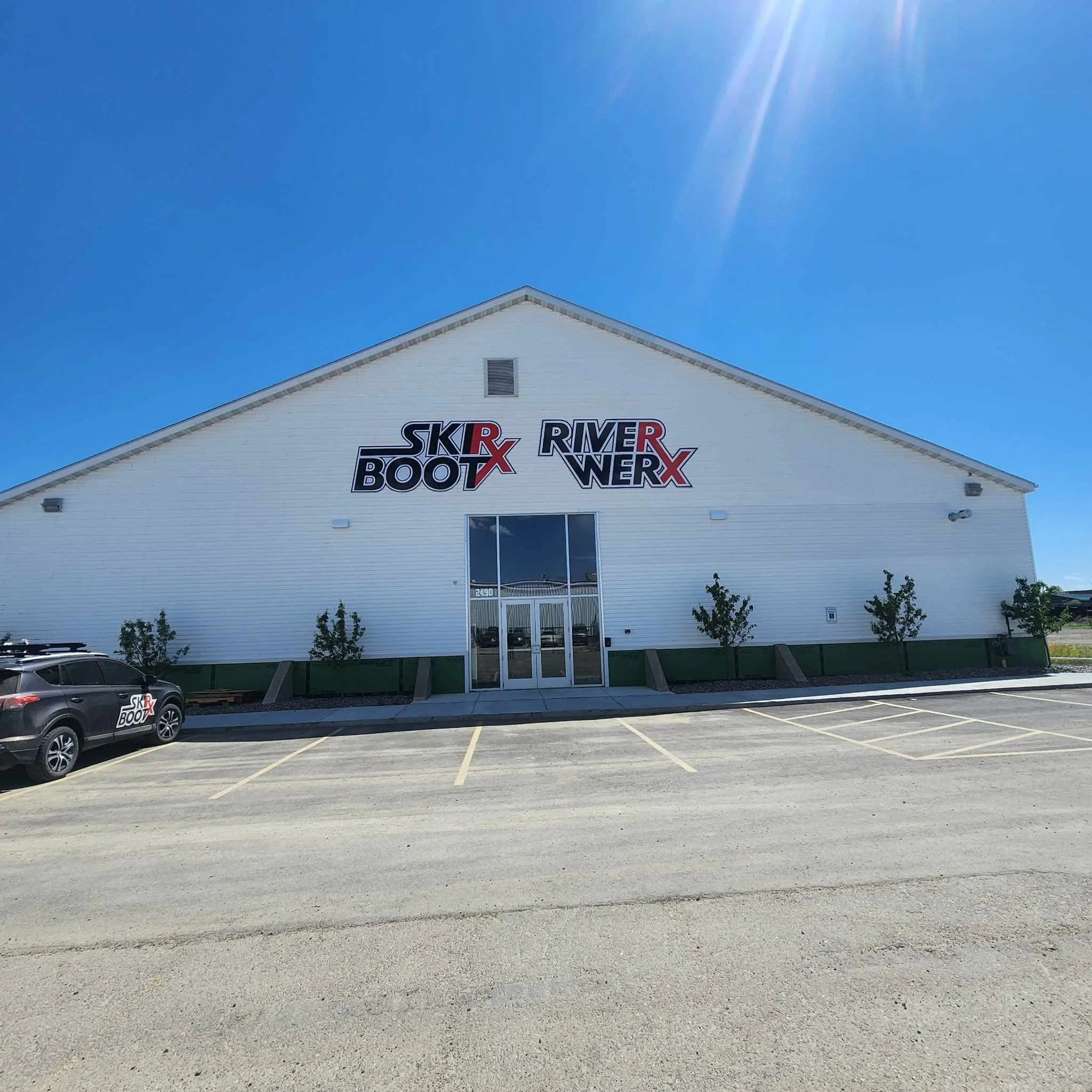 A white building with the signs 'Ski & Boot' and 'River WERX' on the front. There are three small trees and a parking lot with a few cars parked, including a black car with similar signage.