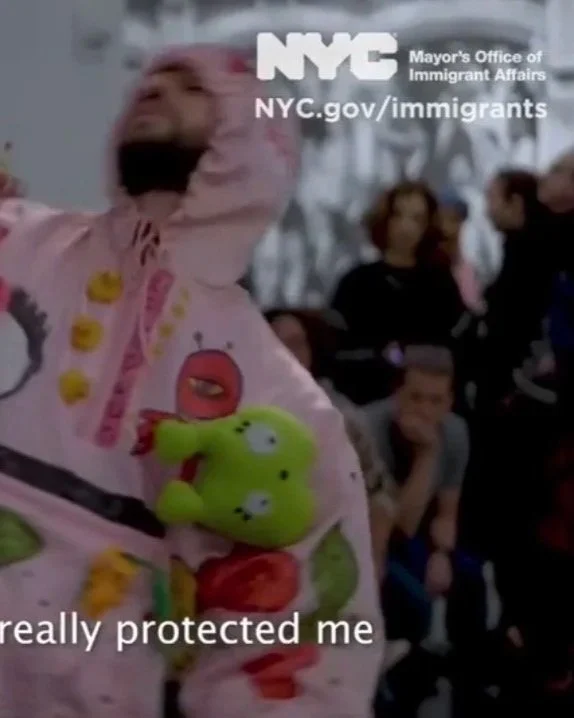 Christopher dances euphorically at the Brooklyn Museum wearing an ornate pink hoodie. A "NYC Mayor's Office of Immigrant Affairs" logo appears in the corner.