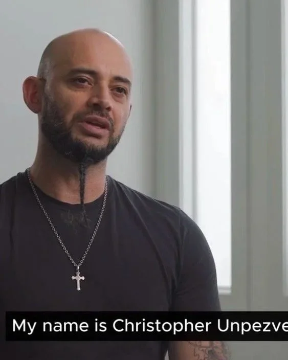 Christopher, wearing a black t-shirt and a silver cross necklace, looks directly into the camera while discussing his choreography.