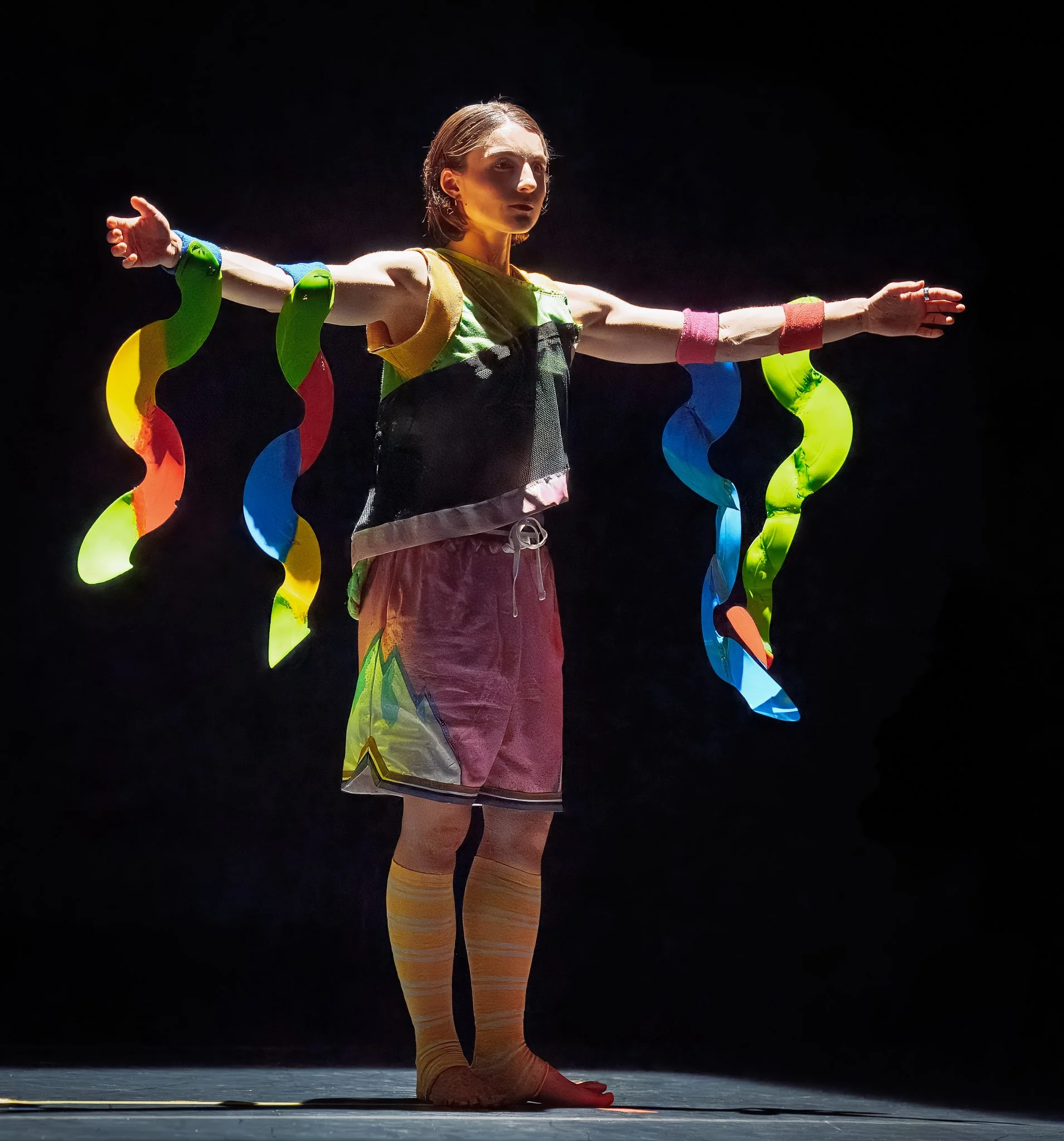 A dancer extends her arms like wings, adorned with vibrant, curved plastic sculptures resembling fish fins. She is illuminated by a warm, ethereal amber light.