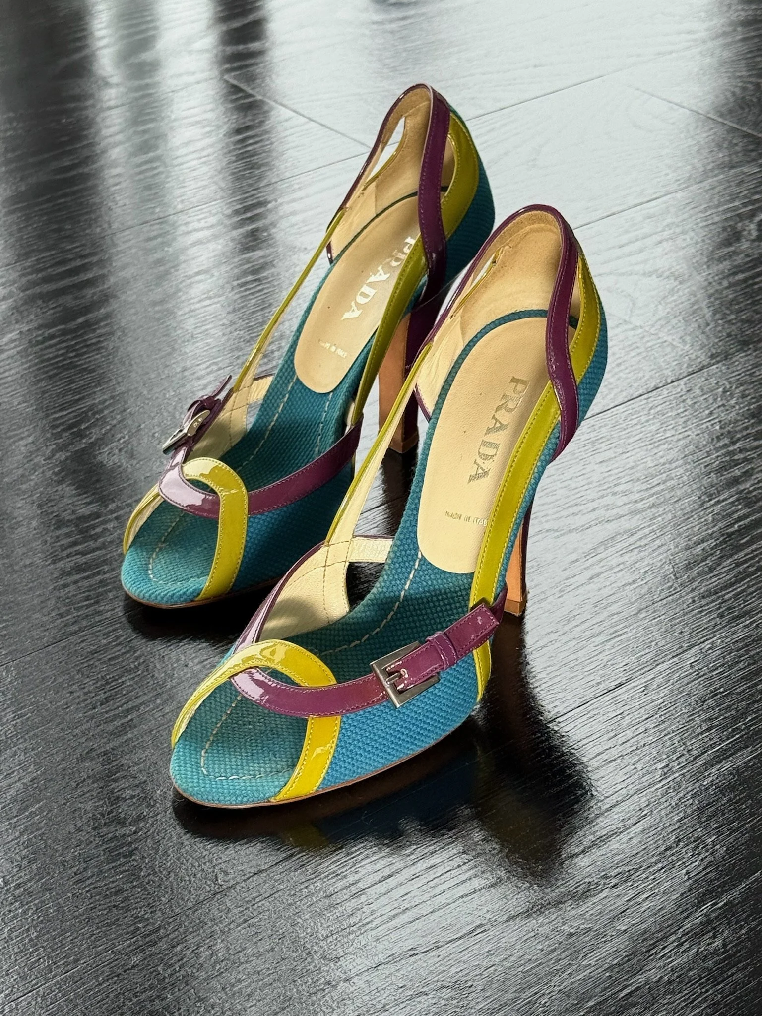 Colorful Prada high-heel shoes with yellow, purple, and teal straps on a dark wooden floor.