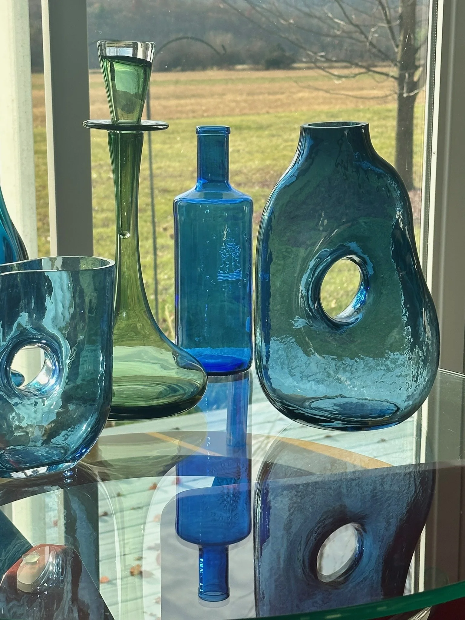 Blue glass vases and bottles on a glass table near a window with a view of a grassy field and trees outside.