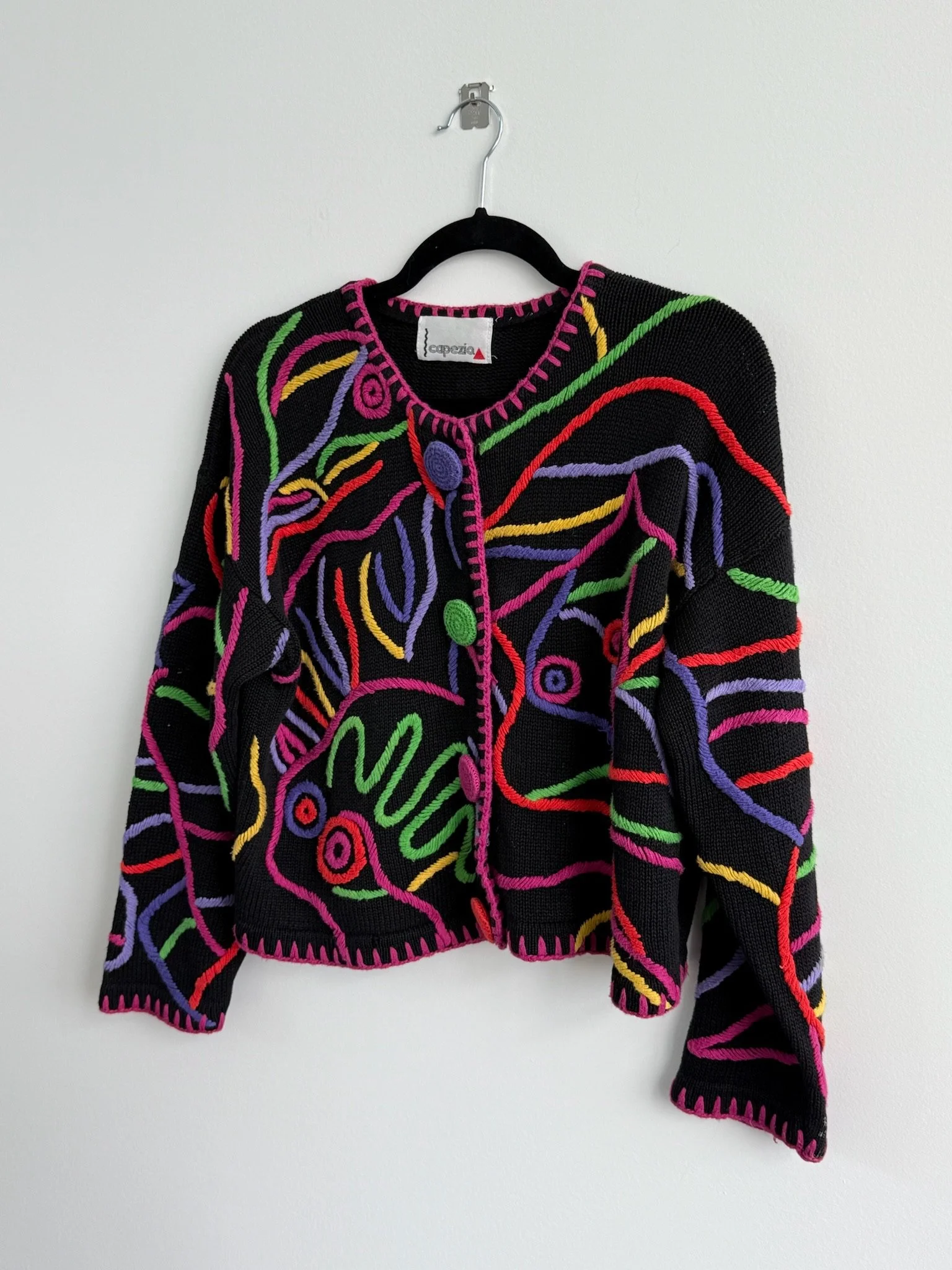 Black cardigan sweater with colorful abstract embroidery, featuring neon pink, green, yellow, purple, and red lines and shapes, hanging on a black hanger against a plain white wall.