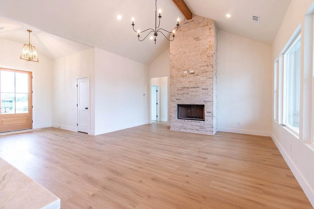 Empty living room with a brick fireplace, large windows, wood flooring, and a high ceiling with a chandelier.