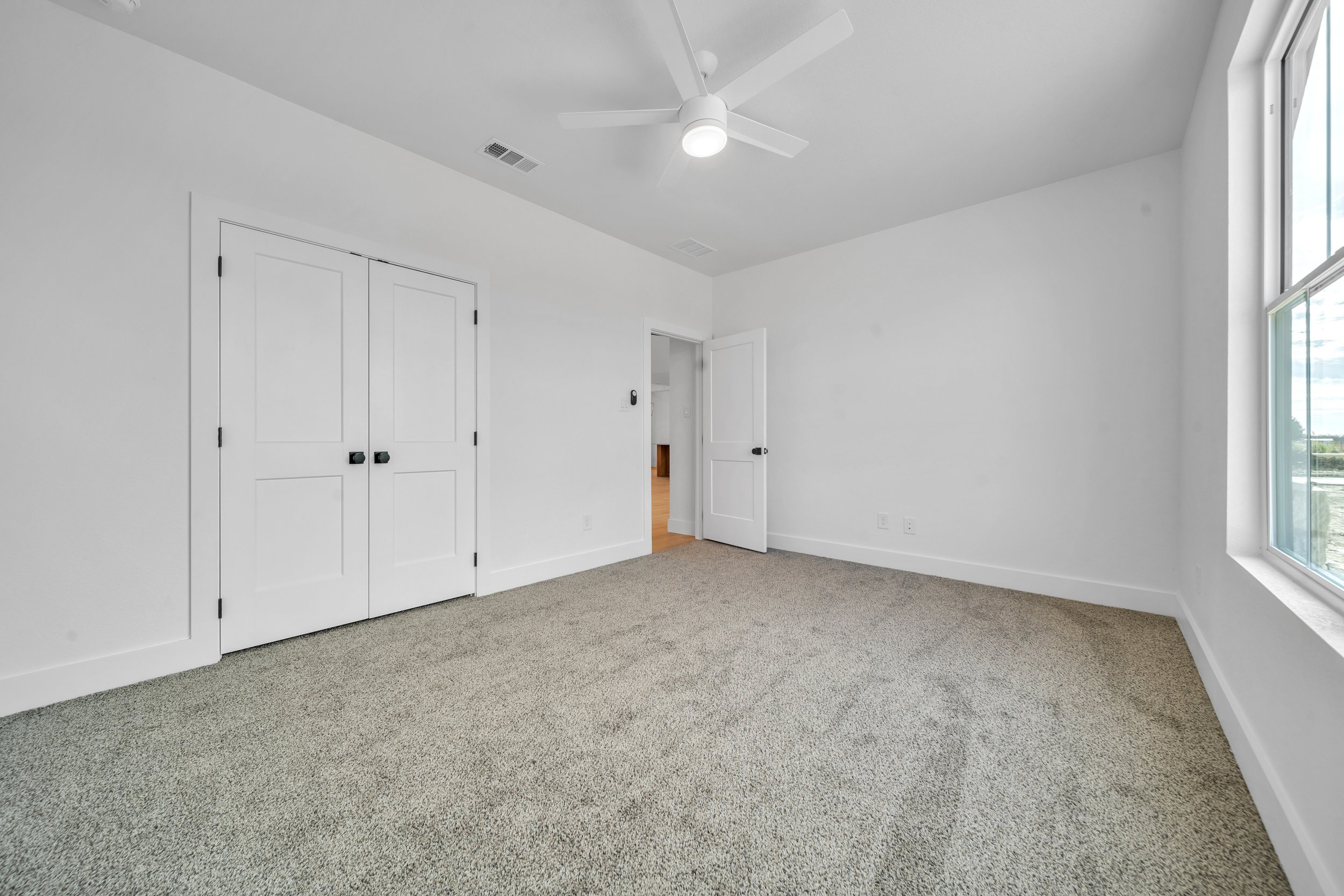 Empty room with white walls, beige carpet, a ceiling fan, a large window, and a closet with double doors.