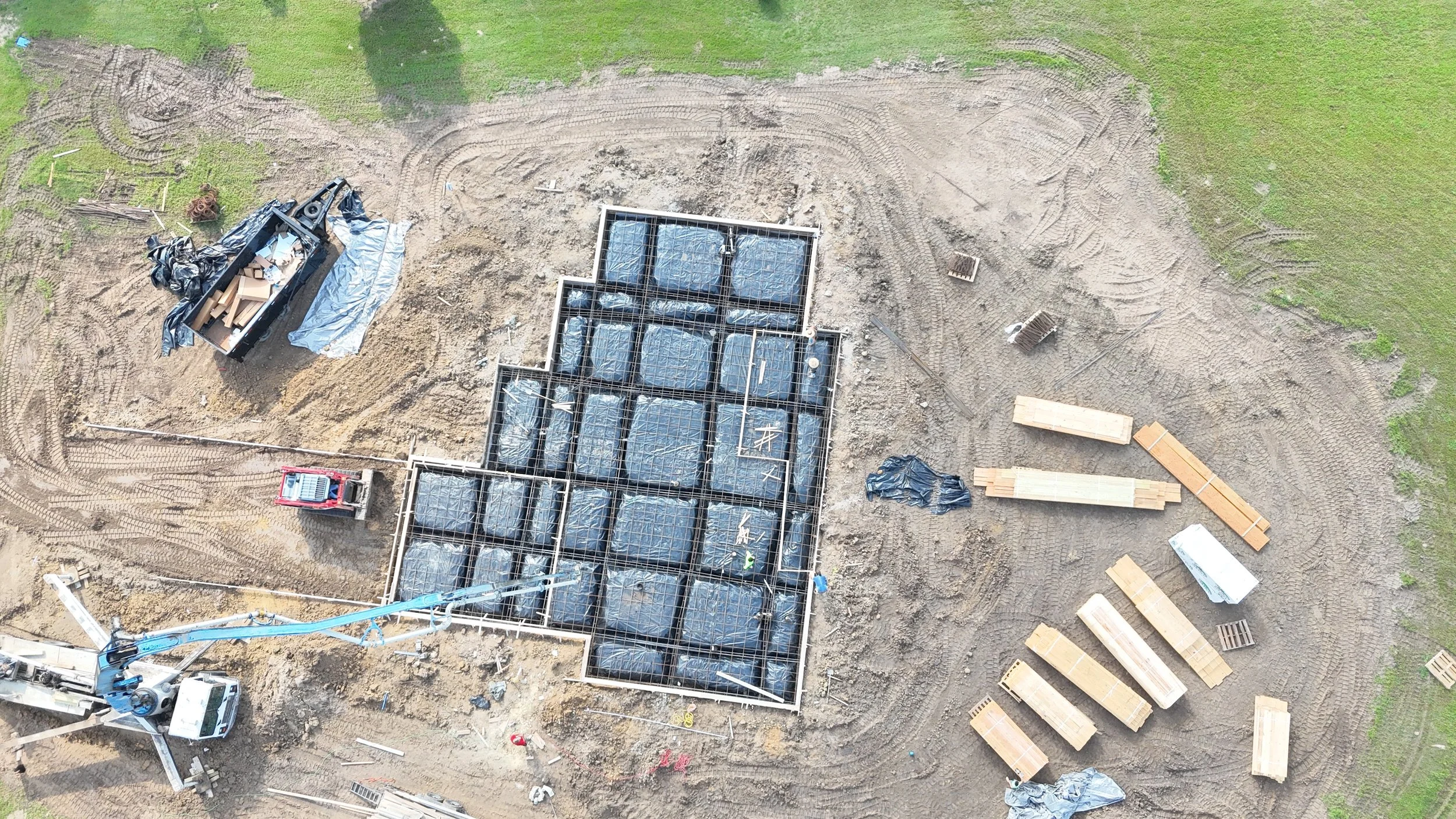 An aerial view of a construction site showing foundation work with metal beams and wrapped materials, construction equipment, and scattered wooden planks on dirt ground surrounded by grass.